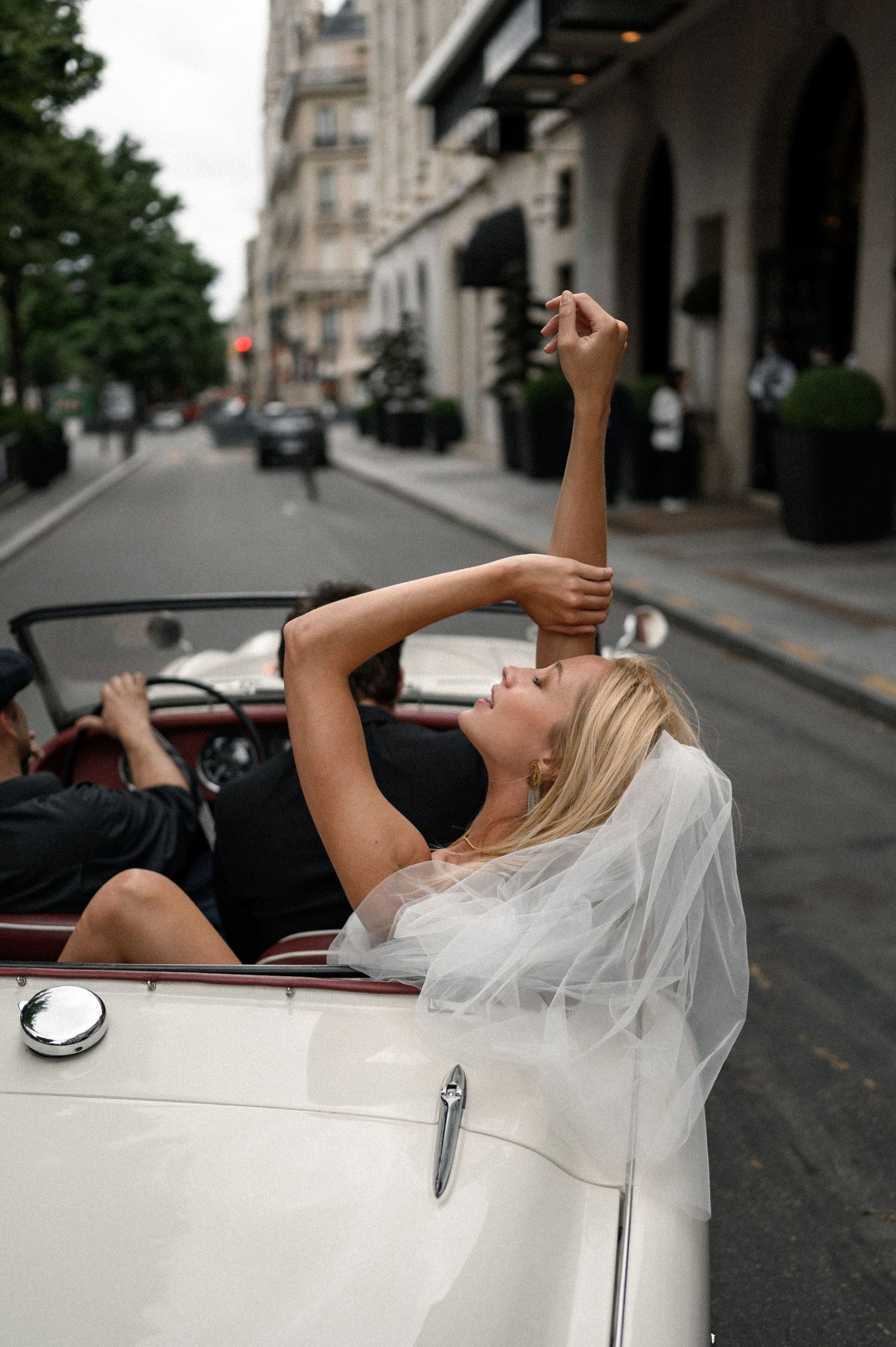 Bride in white veil reclines in vintage white convertible on elegant Paris boulevard, arm raised gracefully.