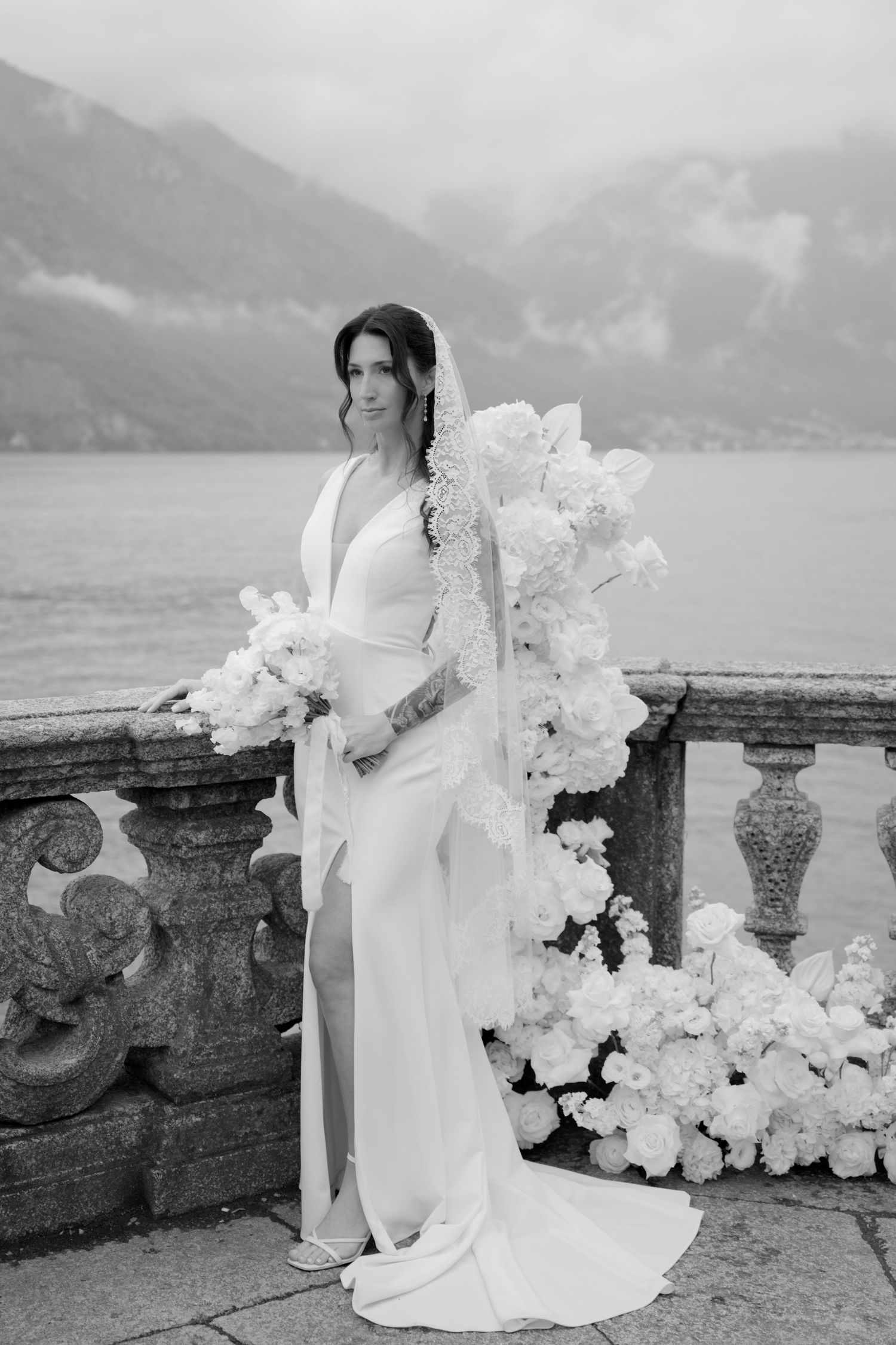 Elegant bride in white gown and floral veil poses by stone balustrade with lake and mountains backdrop.