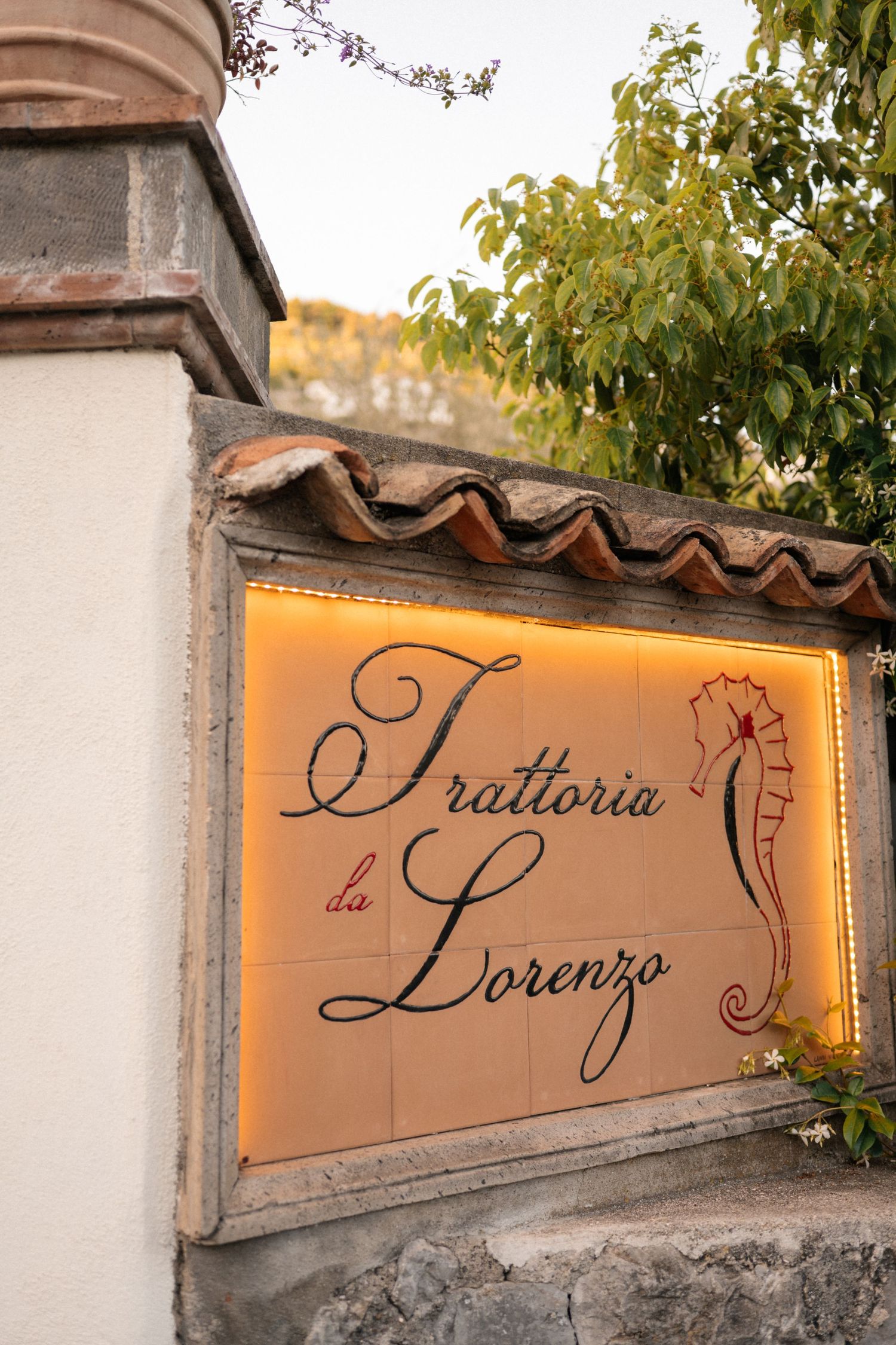 Illuminated tile sign for Trattoria da Lorenzo restaurant with seahorse logo mounted on stone wall.