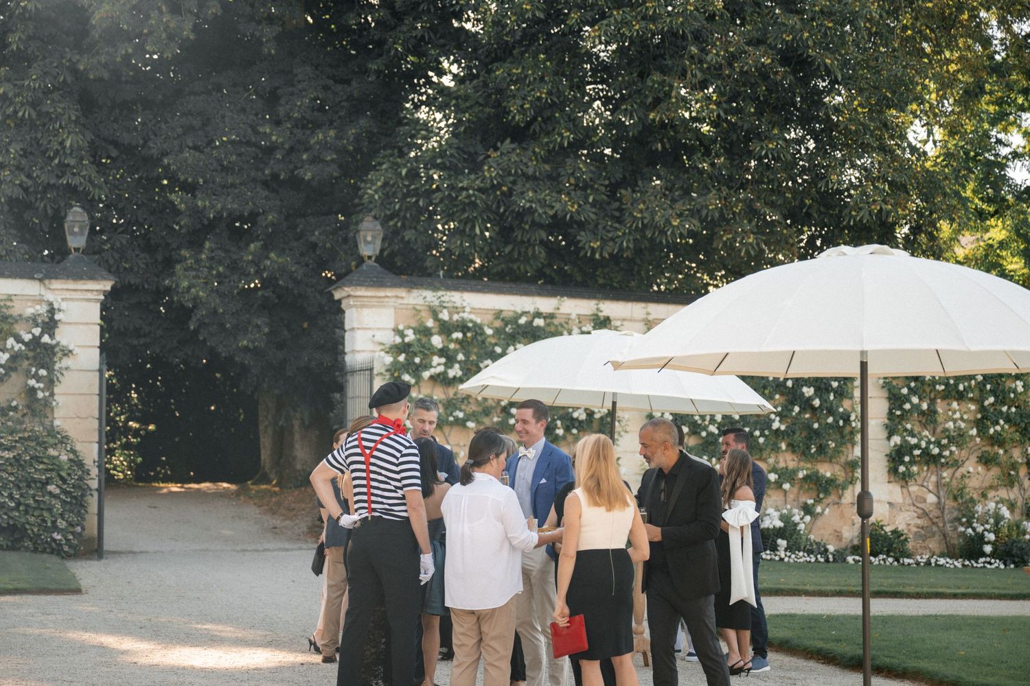 Elegantly dressed guests socializing under white parasols in the lush garden of a French chateau.