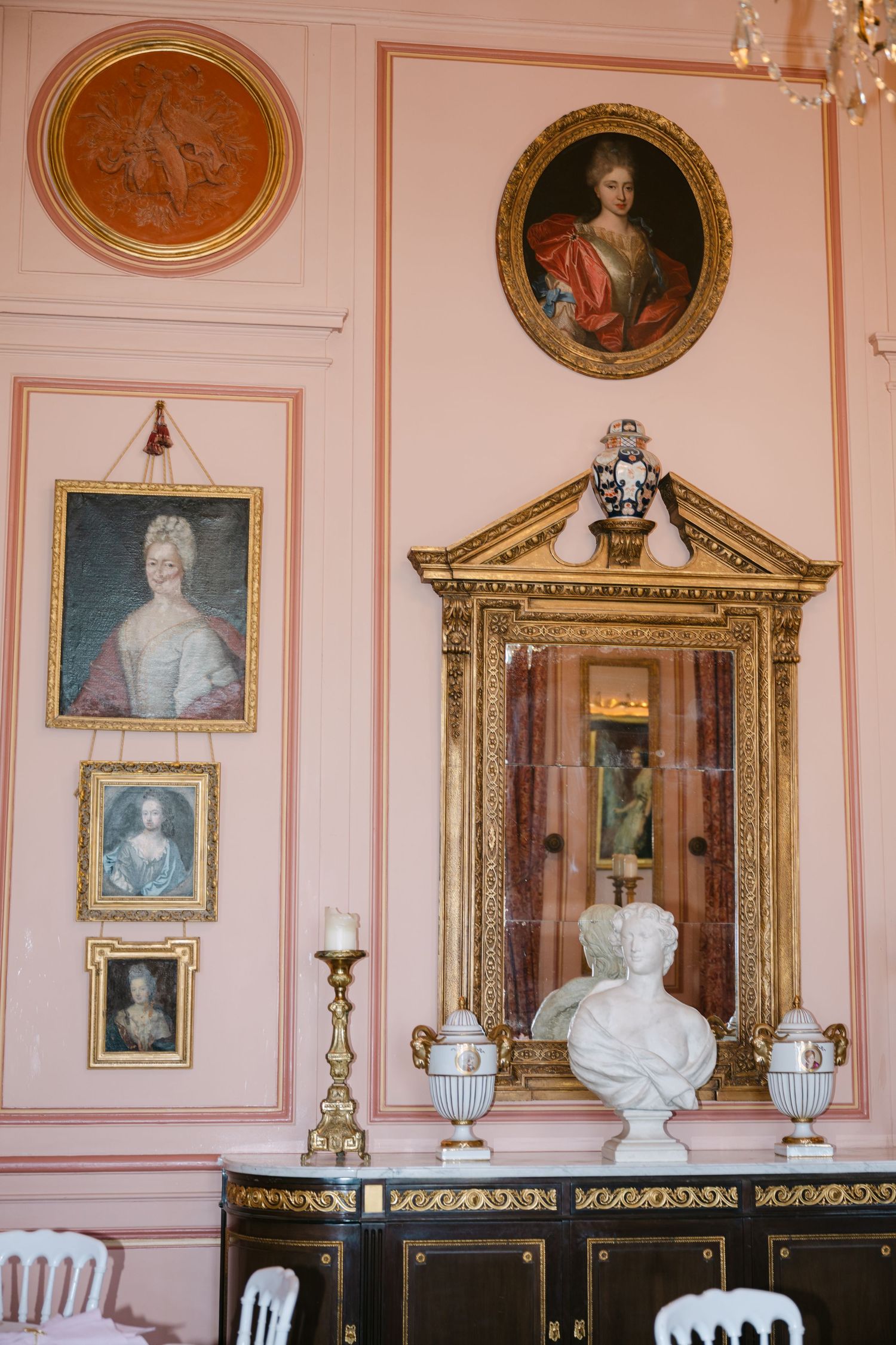 Ornate pink chateau room featuring gilded mirror, marble bust, candelabra, and antique portrait paintings on the wall.