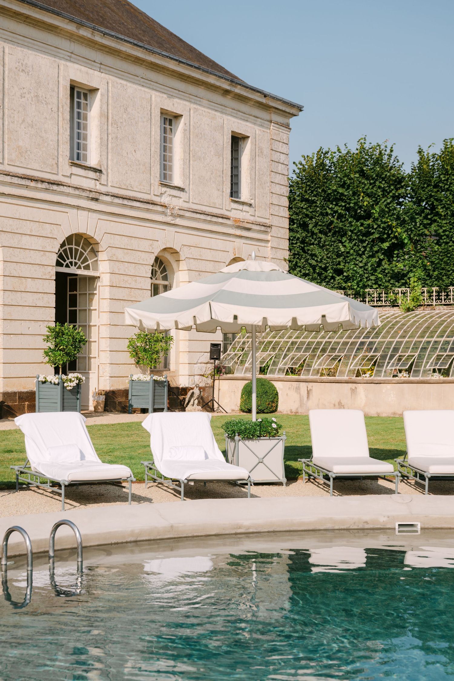 Elegant poolside lounge chairs and striped umbrella beside a classic stone French chateau with manicured gardens.