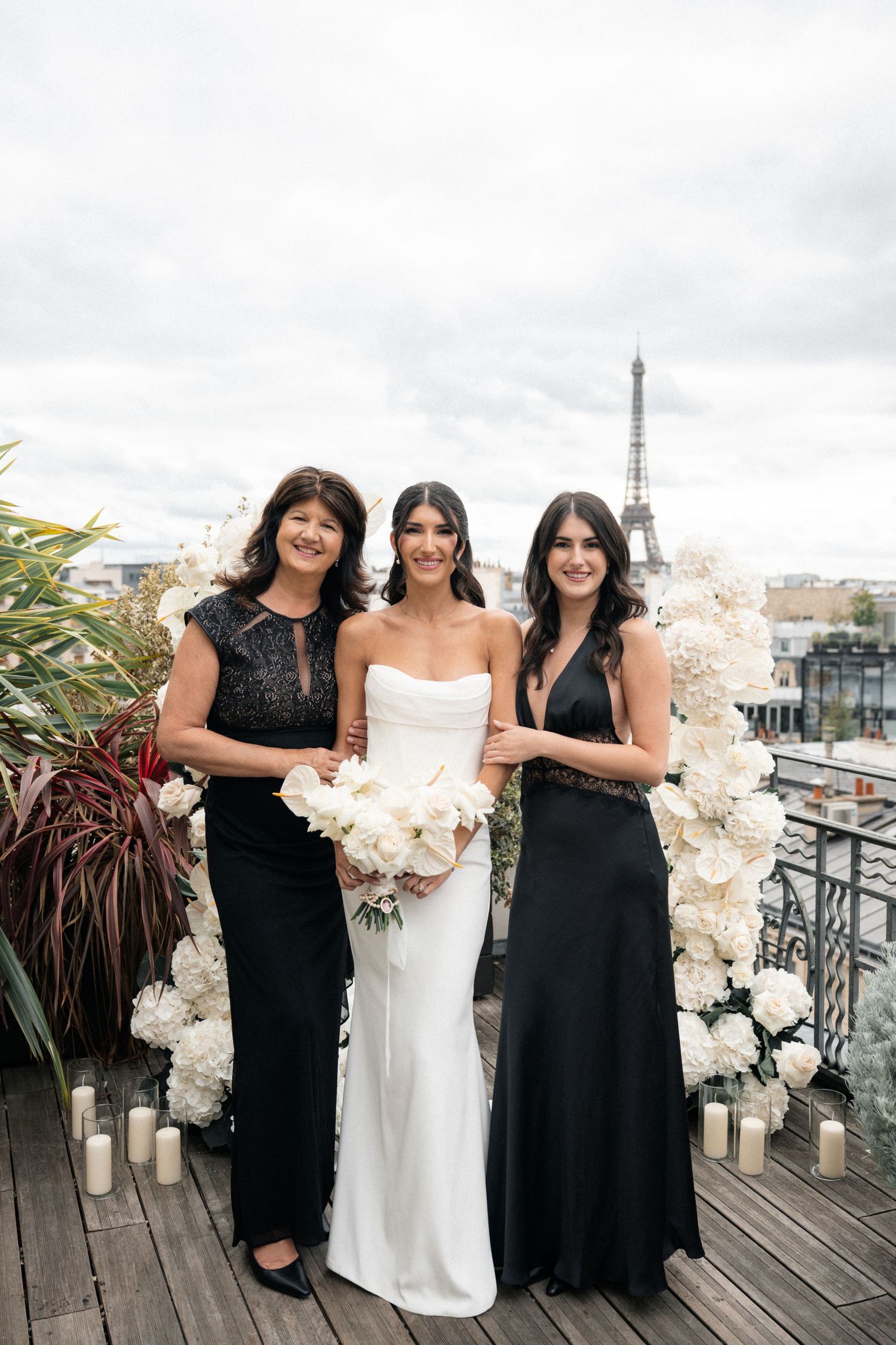 Bride in white gown poses with two women in black dresses on Paris rooftop with Eiffel Tower backdrop.