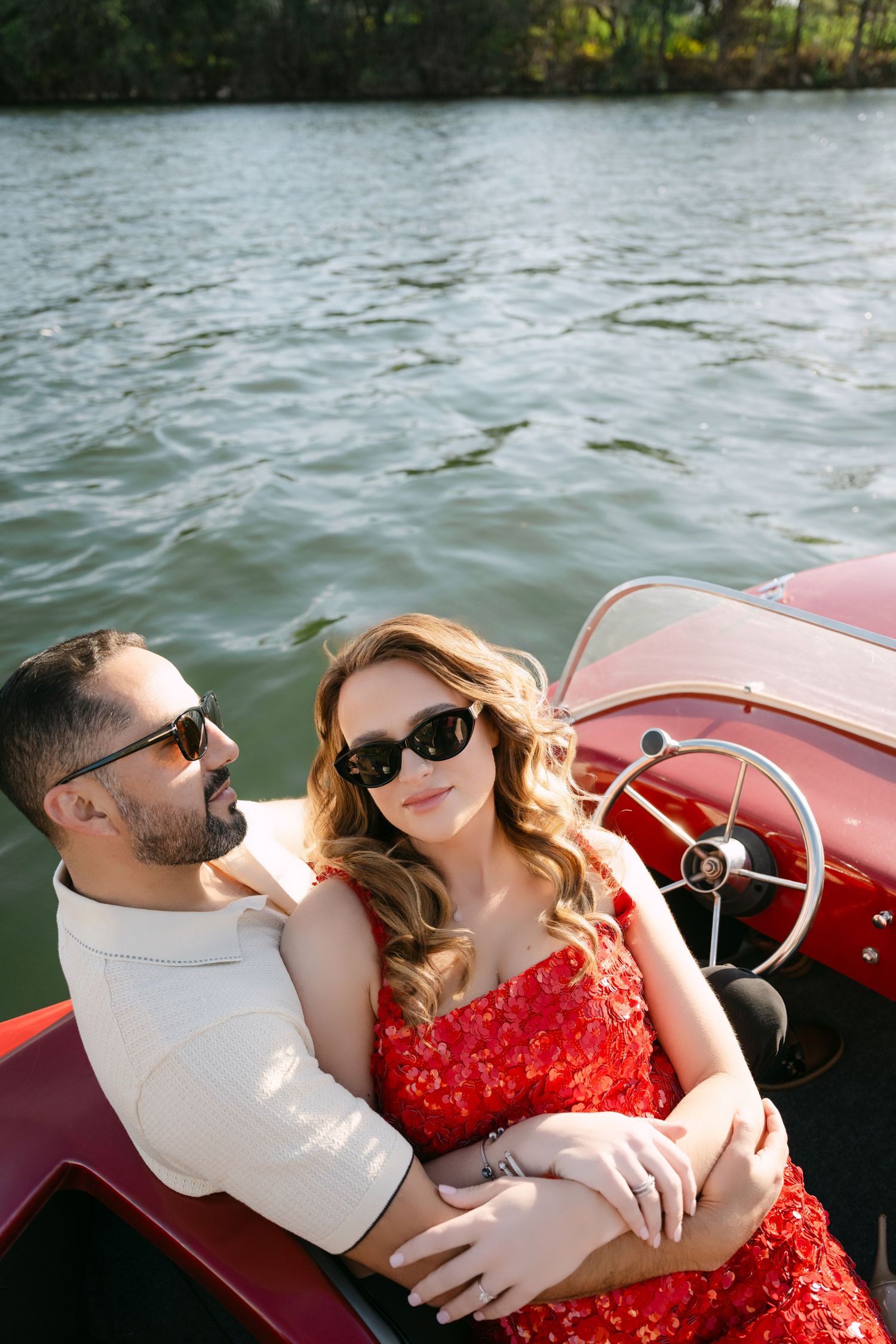 Romantic couple cuddles on classic red wooden boat on calm river, both wearing stylish sunglasses.