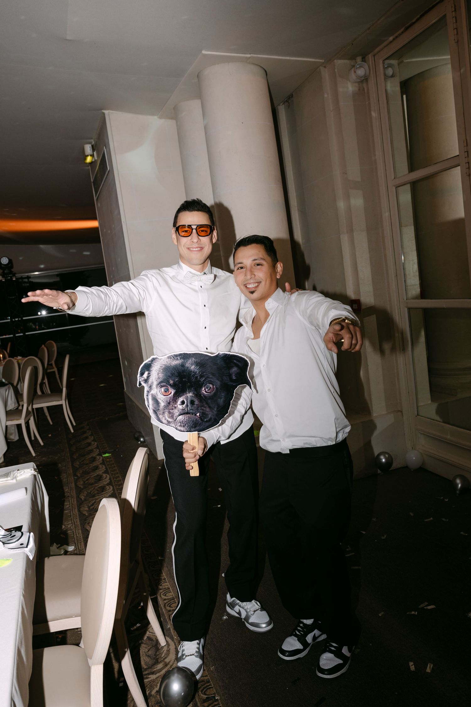 Two men posing with a novelty pug dog face prop on a stick at a party.