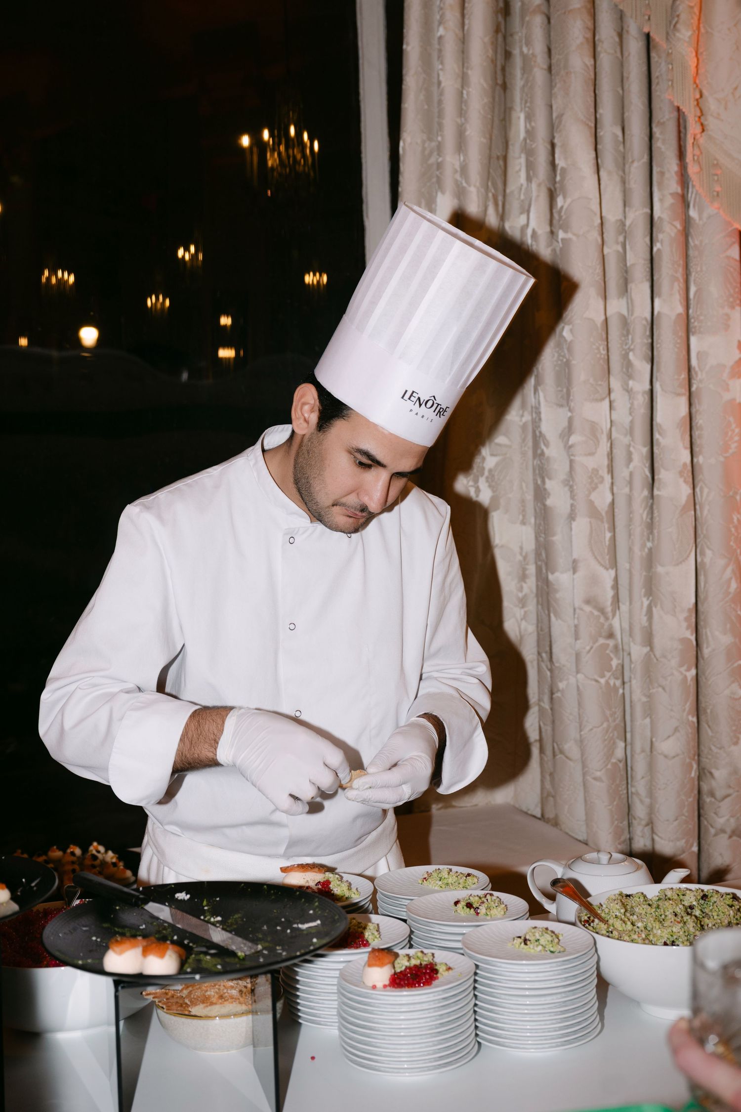Professional chef in white uniform and tall hat plating elegant appetizers at wedding reception.