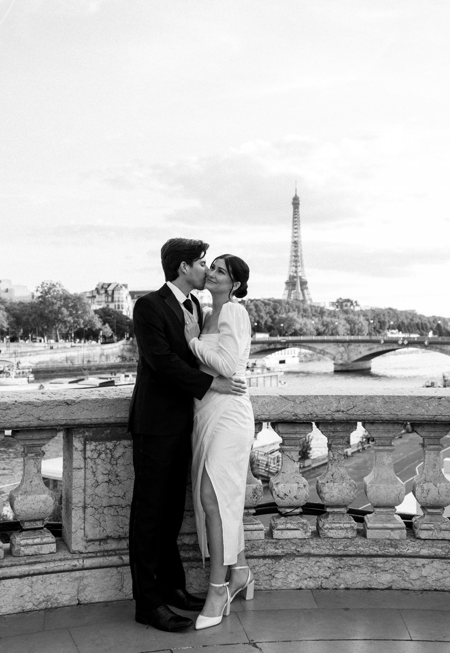 A couple shares a romantic kiss on a bridge with the Eiffel Tower in the background, black and white.