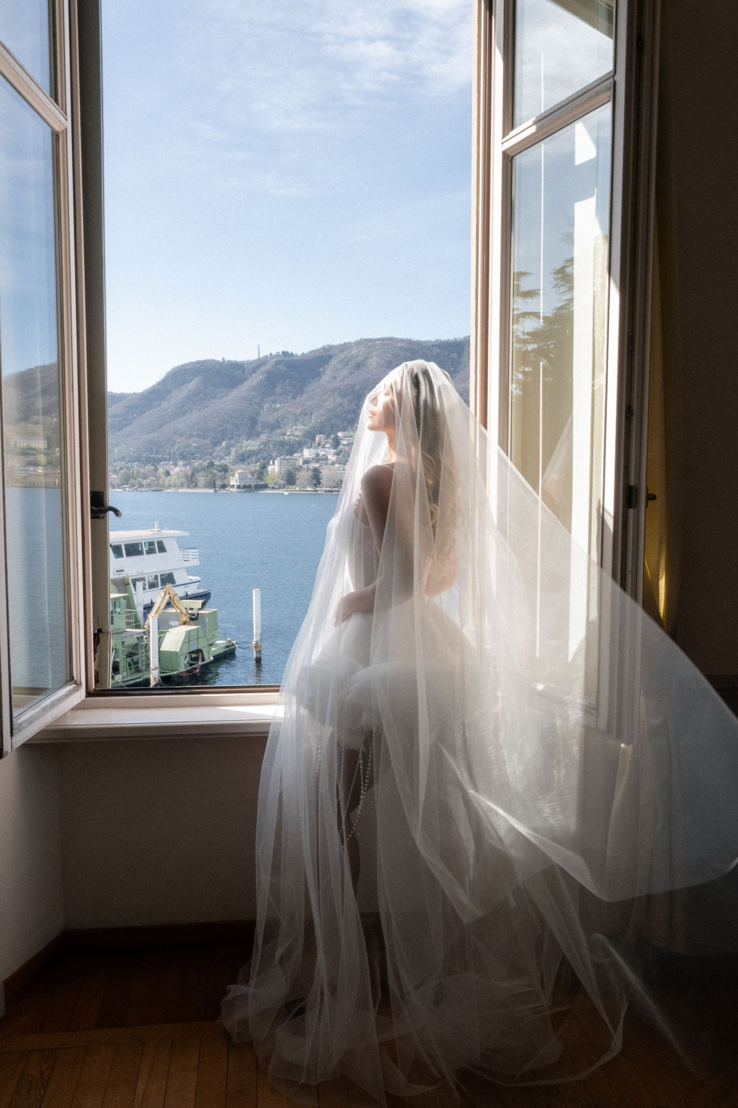 Bride in flowing veil standing at open window overlooking Lake Como with mountains and ferry in background.