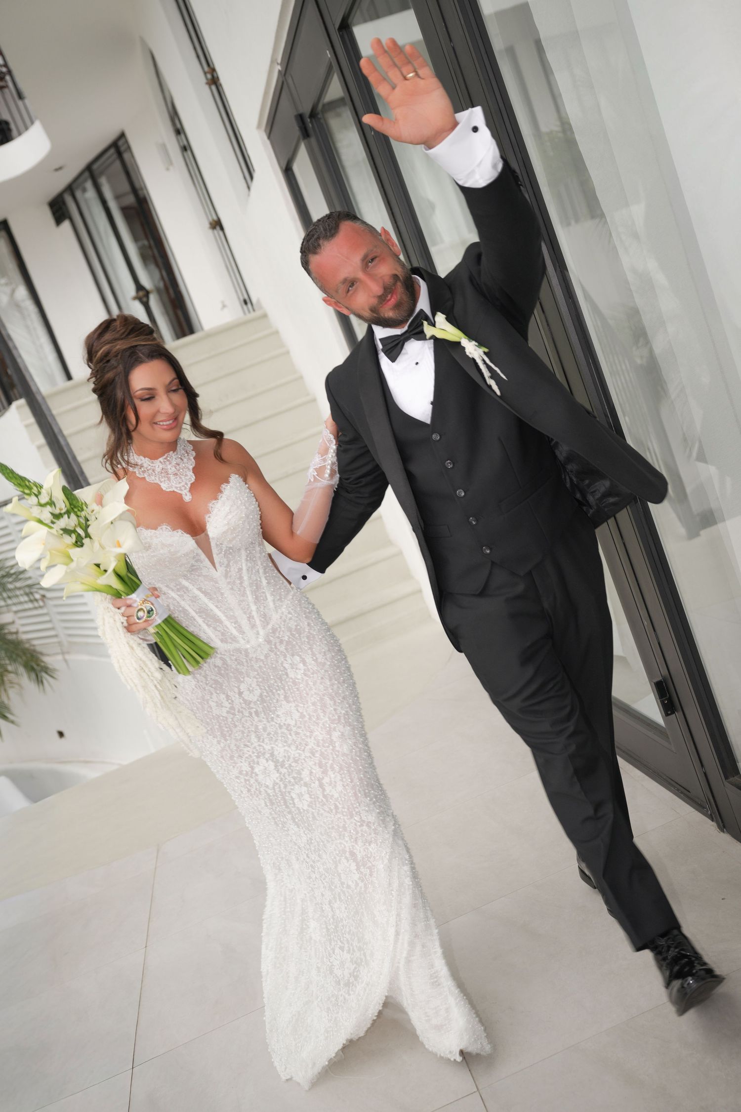 Bride in lace gown holding bouquet walks with tuxedo-clad groom waving at elegant wedding venue.