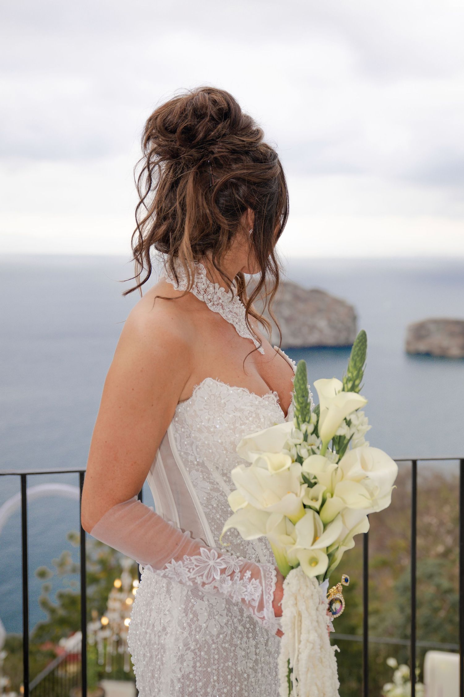 Bride in white lace gown holds white calla lily bouquet overlooking the sea on a cloudy day.