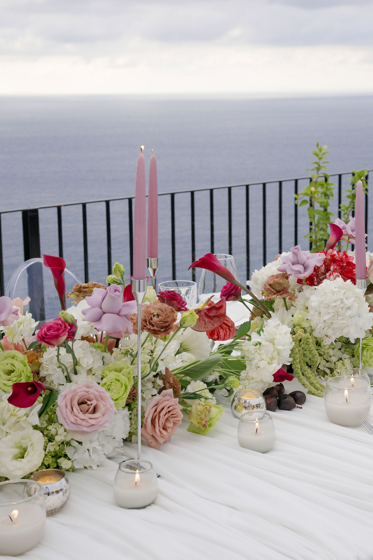 Romantic outdoor oceanview wedding table with lush floral arrangements, pink taper candles, and glowing votive candles.