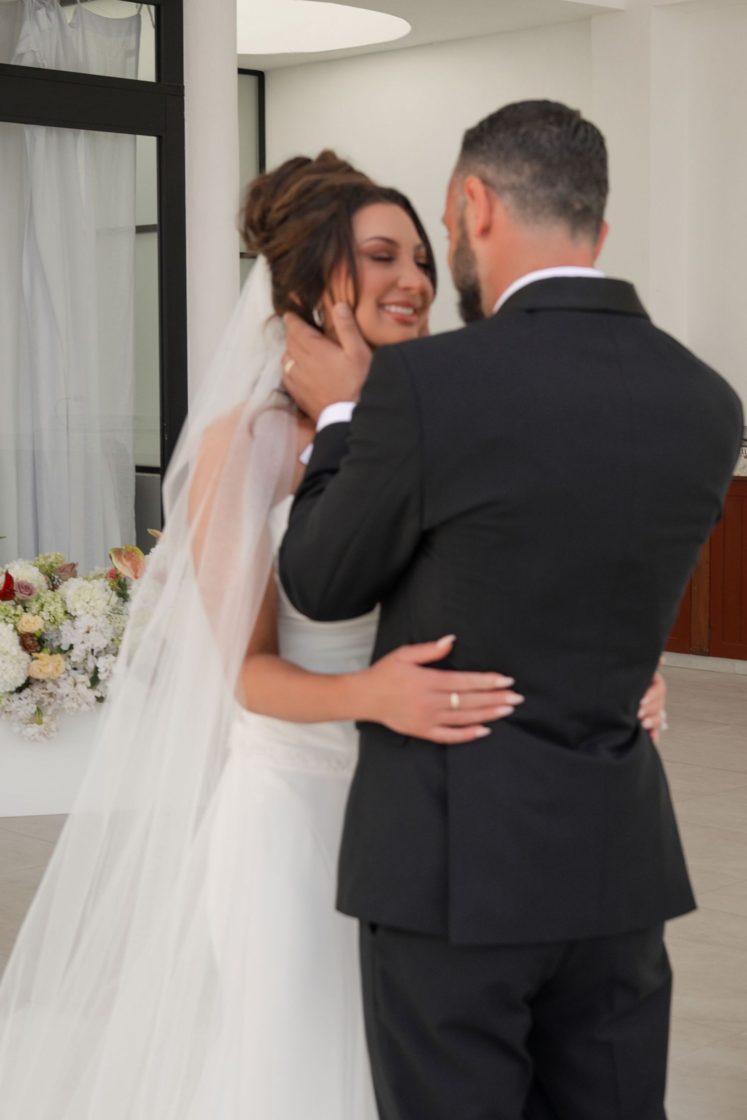 A bride in a white gown and veil smiles as the groom in a black suit tenderly holds her face during their wedding.