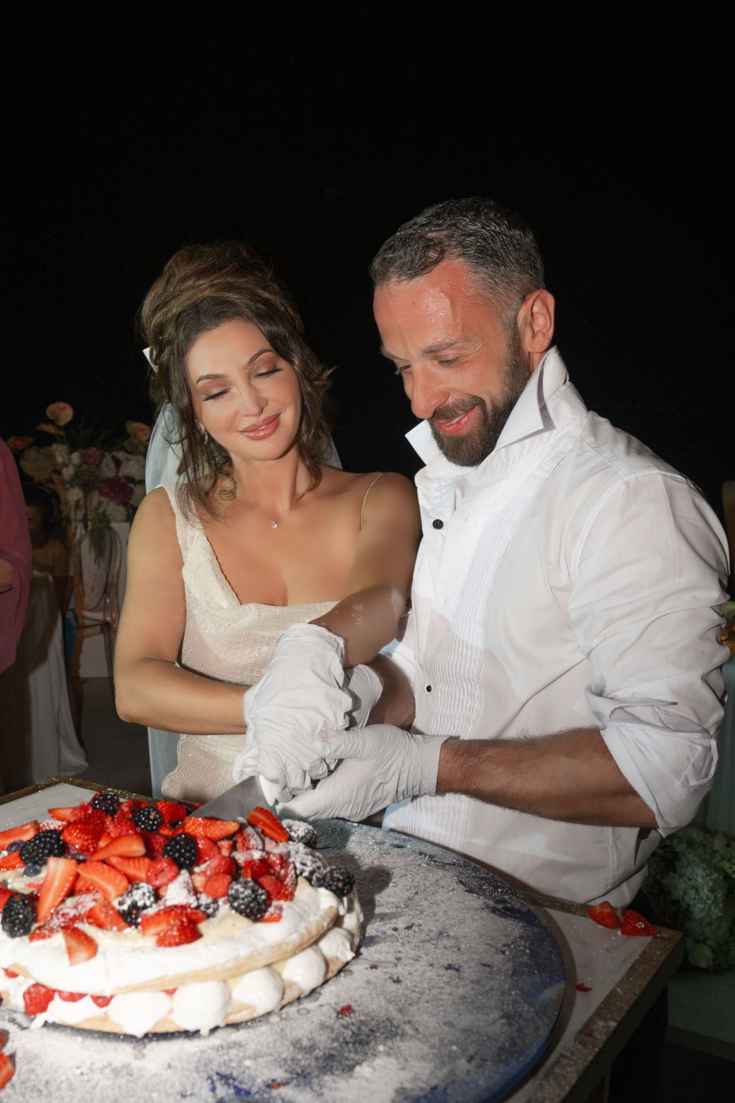 Smiling bride and groom in white gloves cutting a beautiful fresh fruit wedding cake topped with strawberries and blackberries.