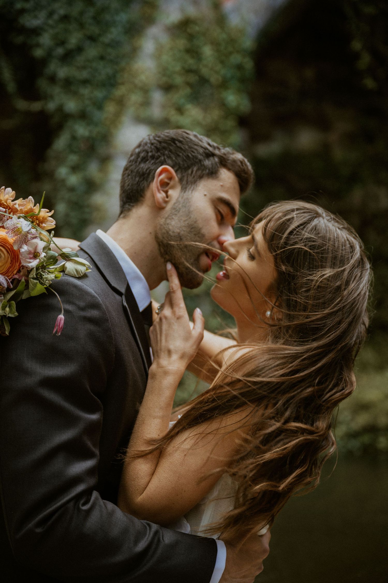Wedding couple nearly kissing outdoors, bride holding vibrant bouquet against lush green ivy backdrop.