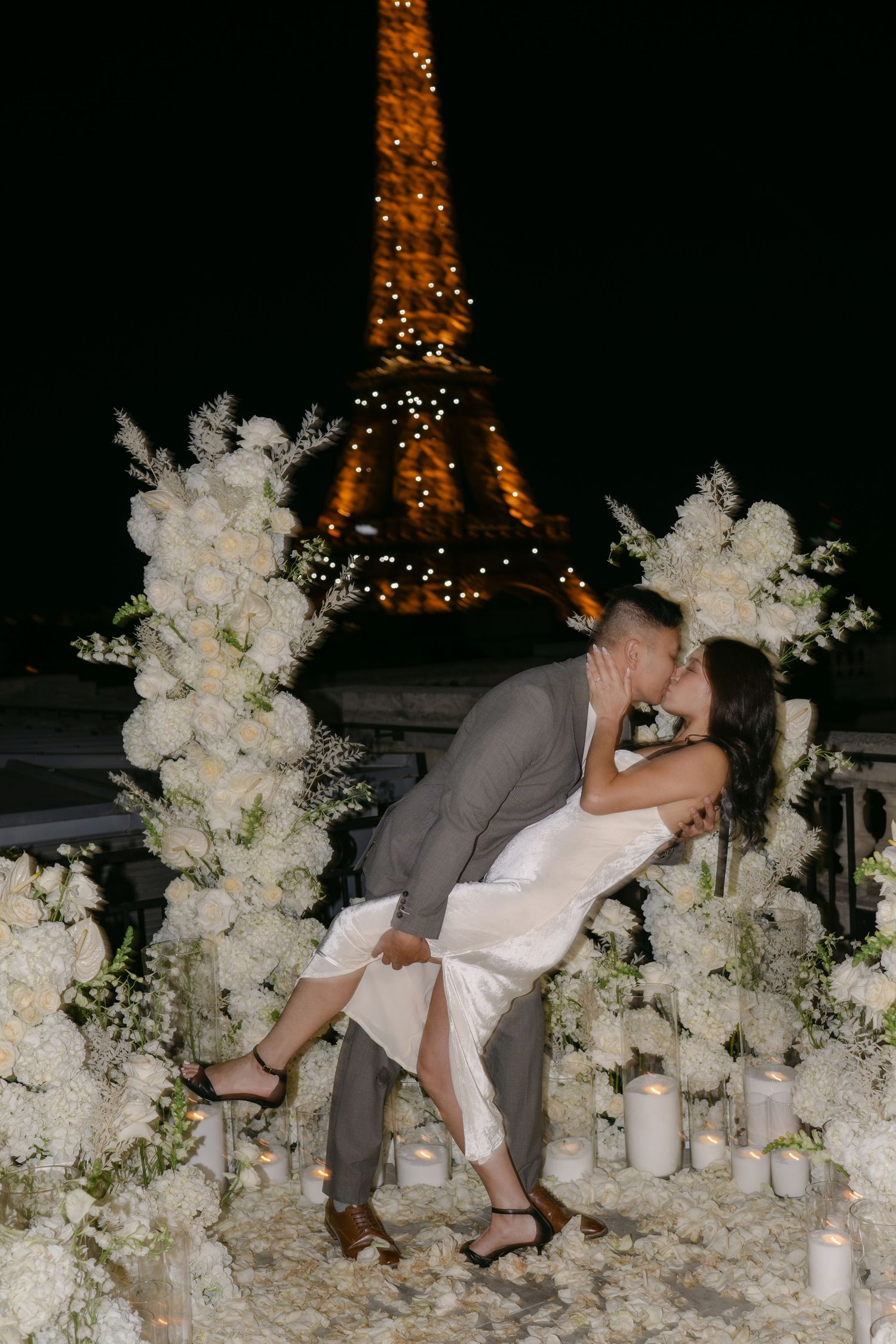 Couple shares a dramatic dip kiss surrounded by white flowers and candles with the illuminated Eiffel Tower at night.