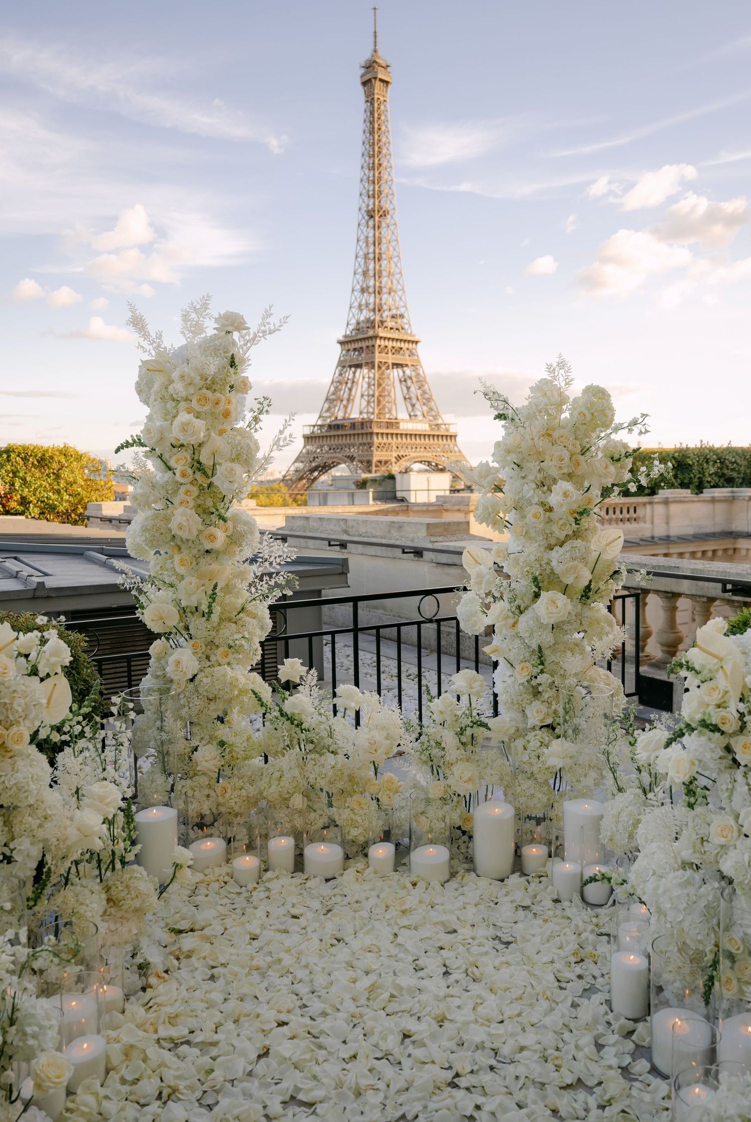 Rooftop proposal setup with white floral arches, rose petals and candles overlooking the Eiffel Tower in Paris at sunset.