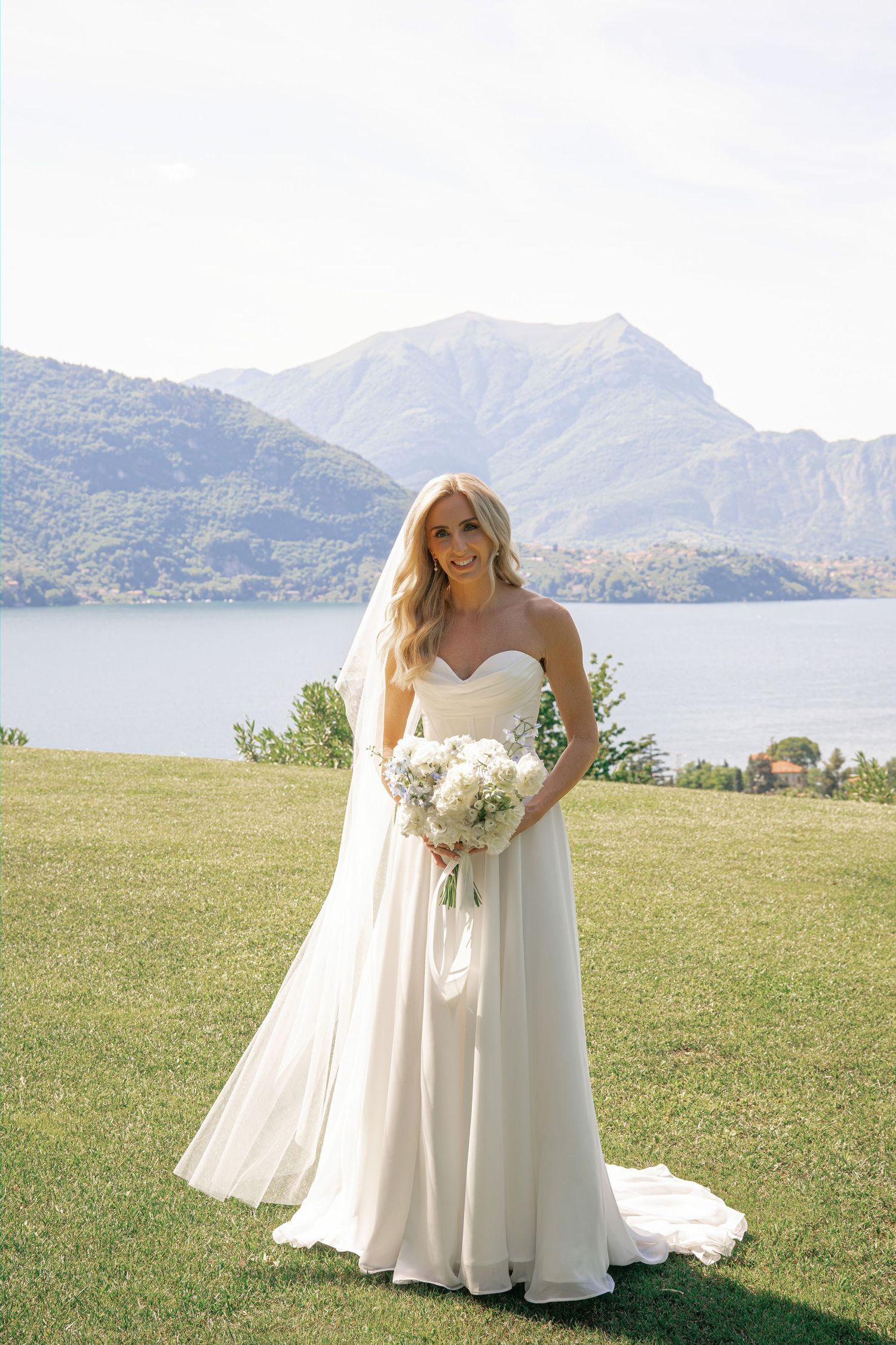 Bride in strapless white gown holding bouquet stands on green lawn with mountain lake backdrop.