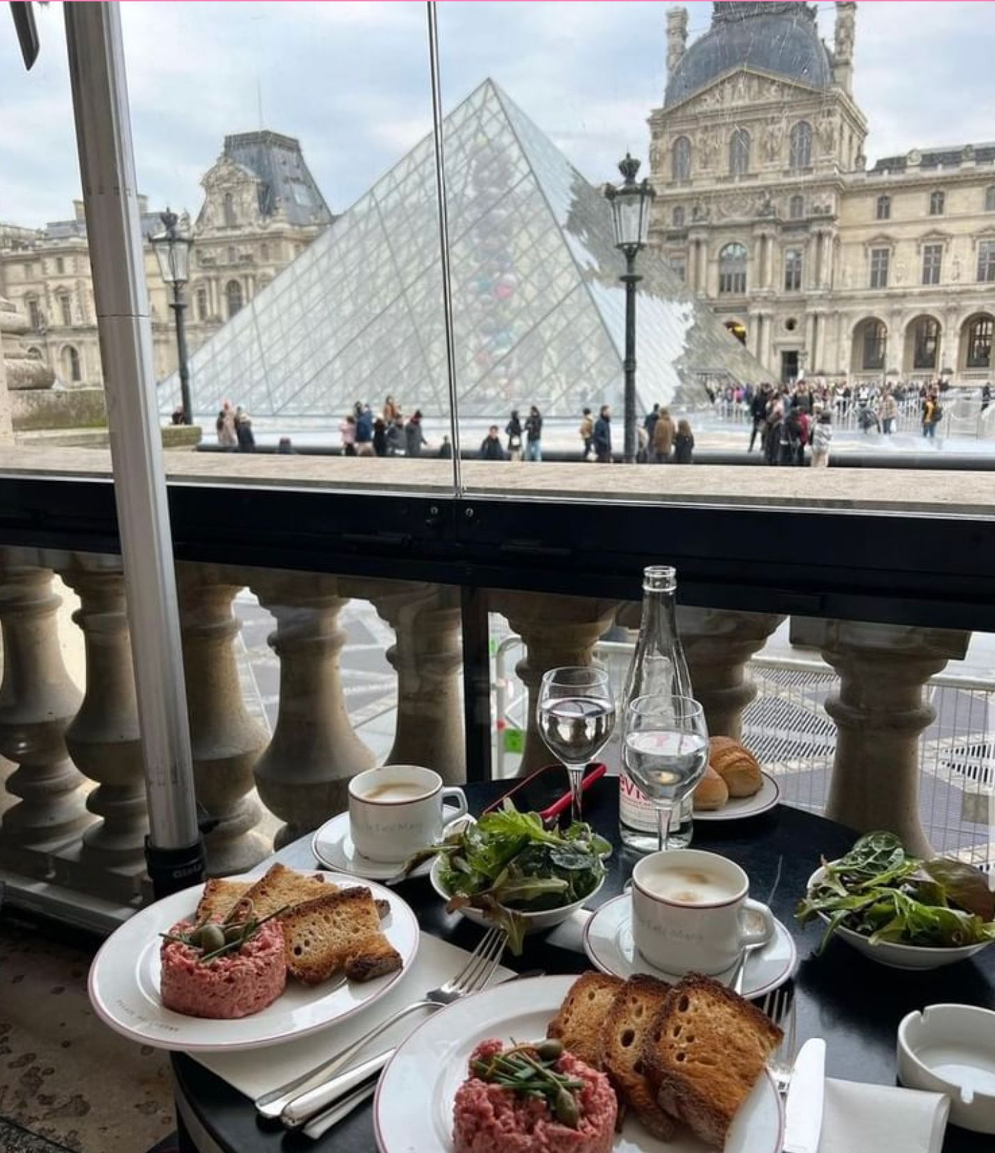 Luxury dining view of the Louvre pyramid with steak tartare and wine on table.