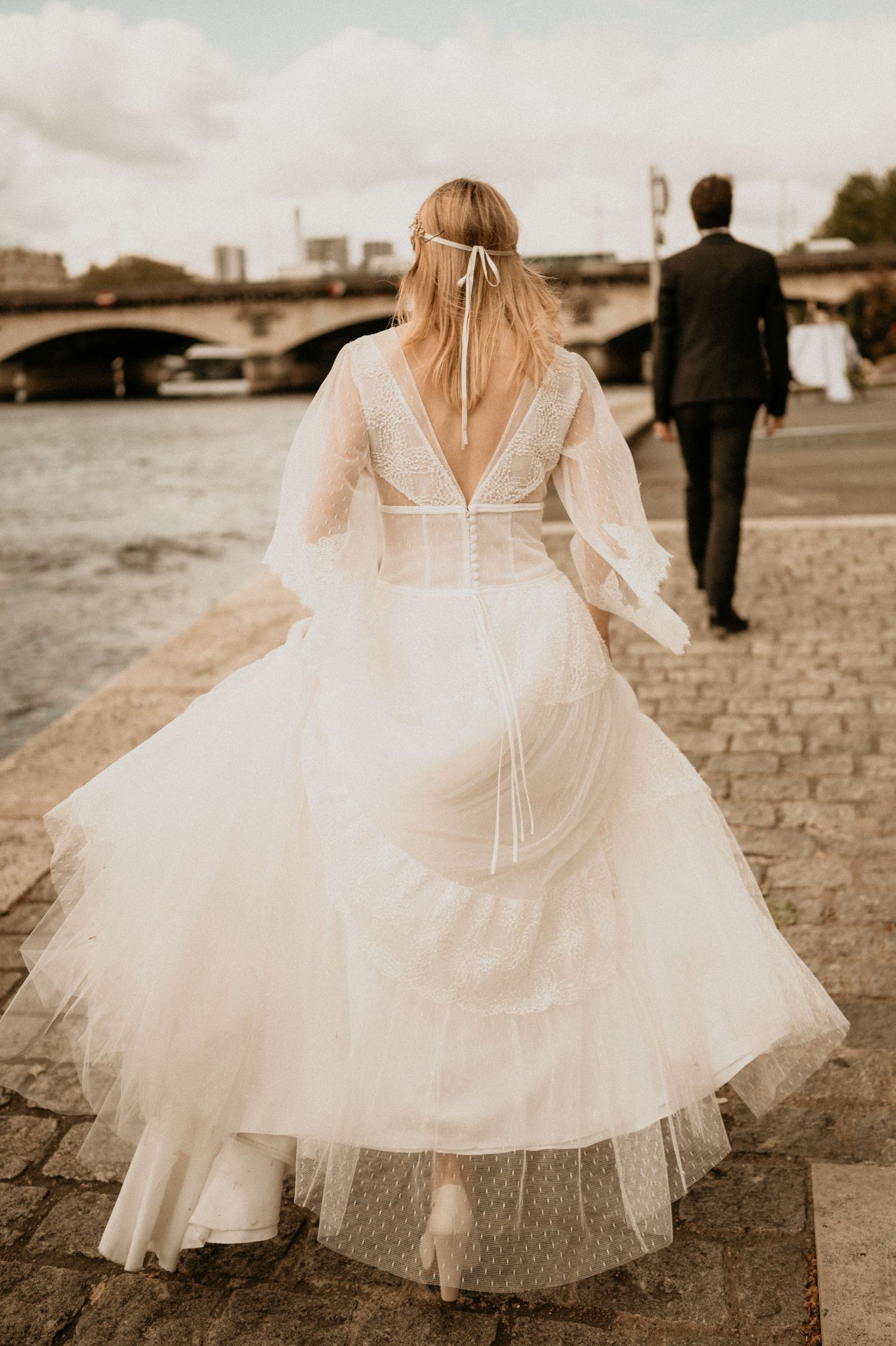 A bride in a flowing white dress walks along the Seine River in Paris with her back to the camera.