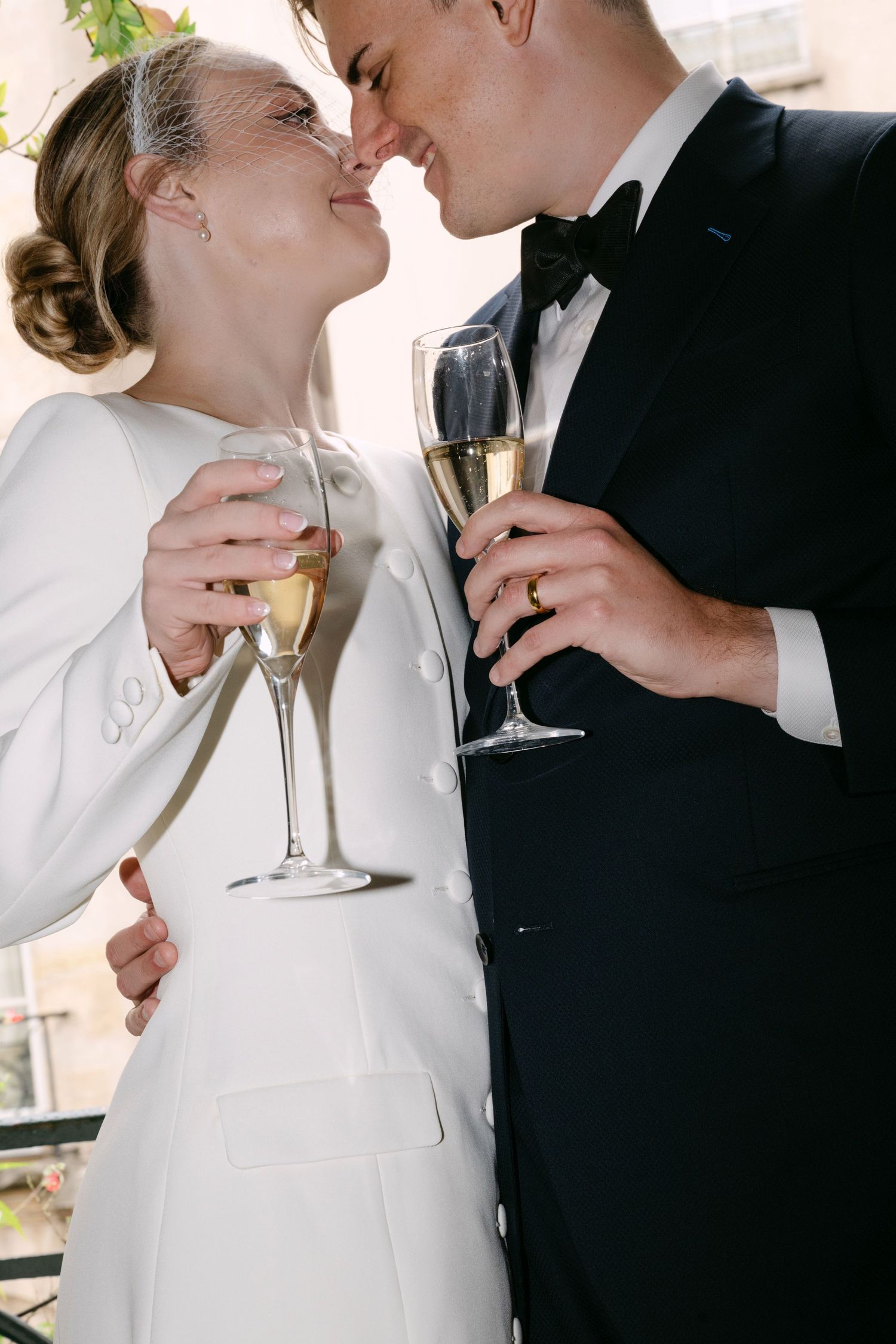 A bride in white and groom in black share champagne while embracing in a romantic moment at their wedding celebration.