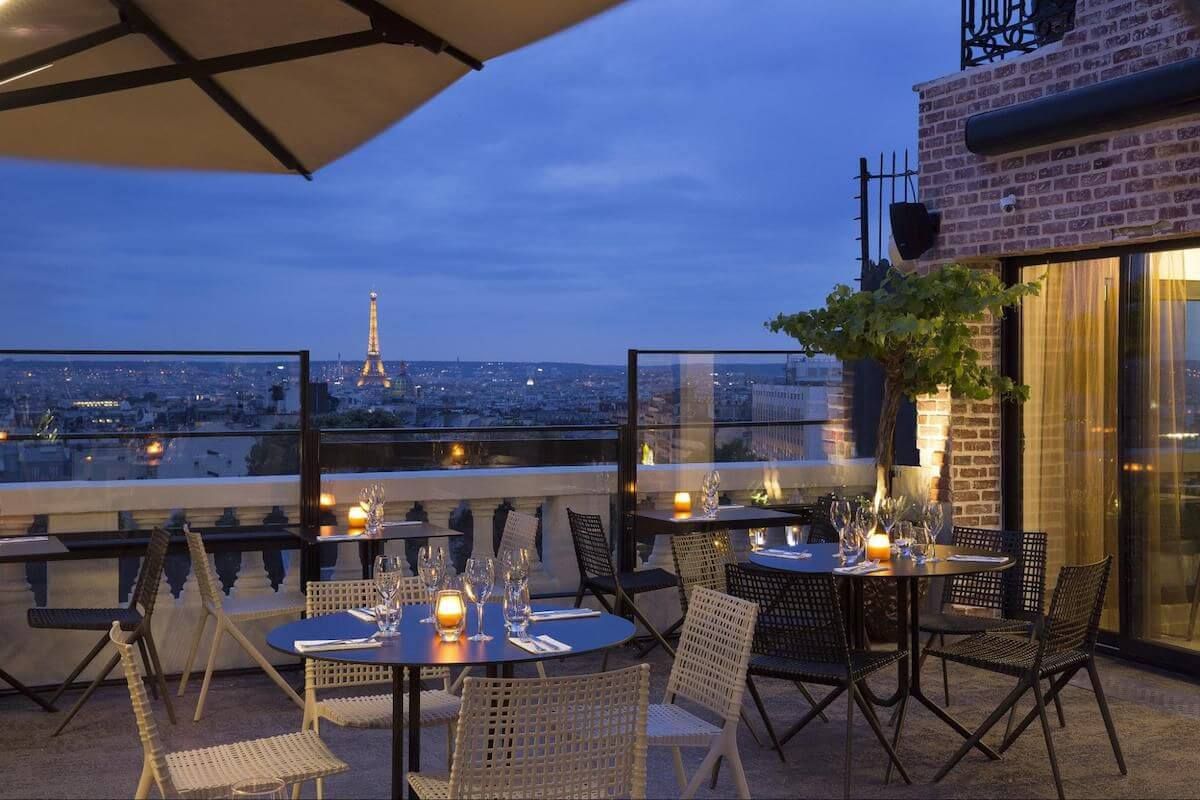 Evening rooftop terrace in Paris with bistro tables and Eiffel Tower glowing in the distance.