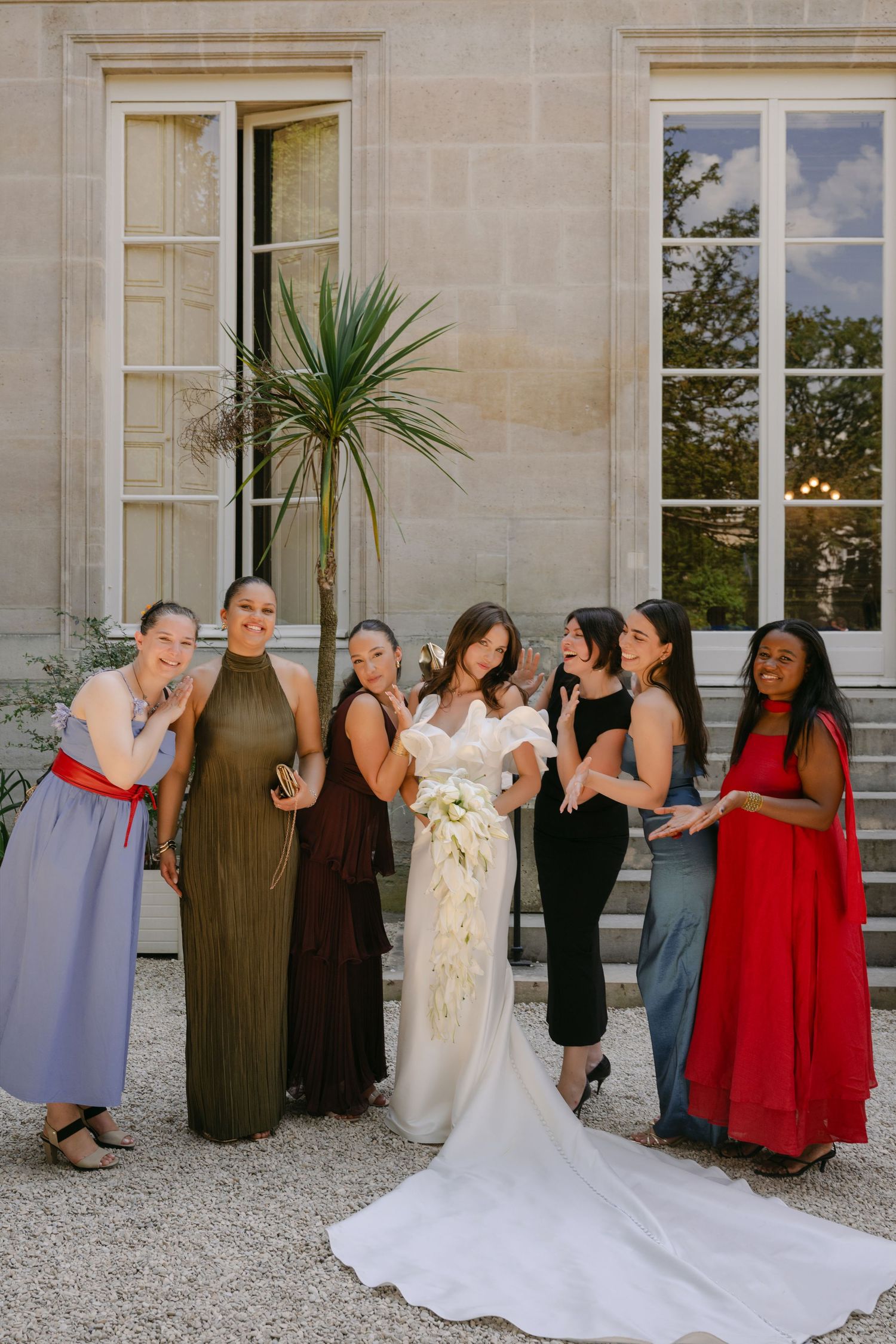 A bride in a white gown poses with six bridesmaids in colorful dresses outside an elegant stone building.