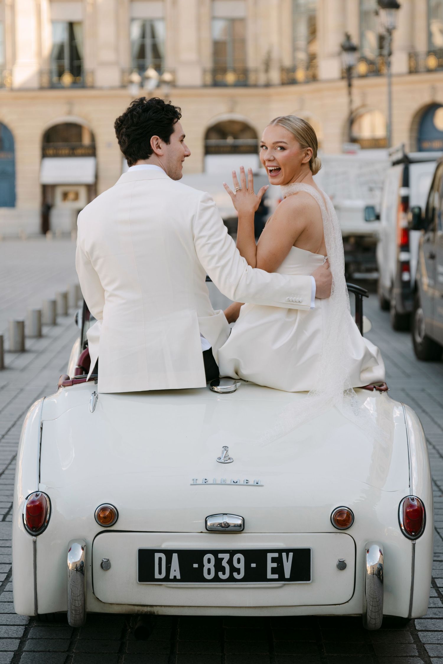 Bride and groom ride a vintage white convertible through a Parisian city square.