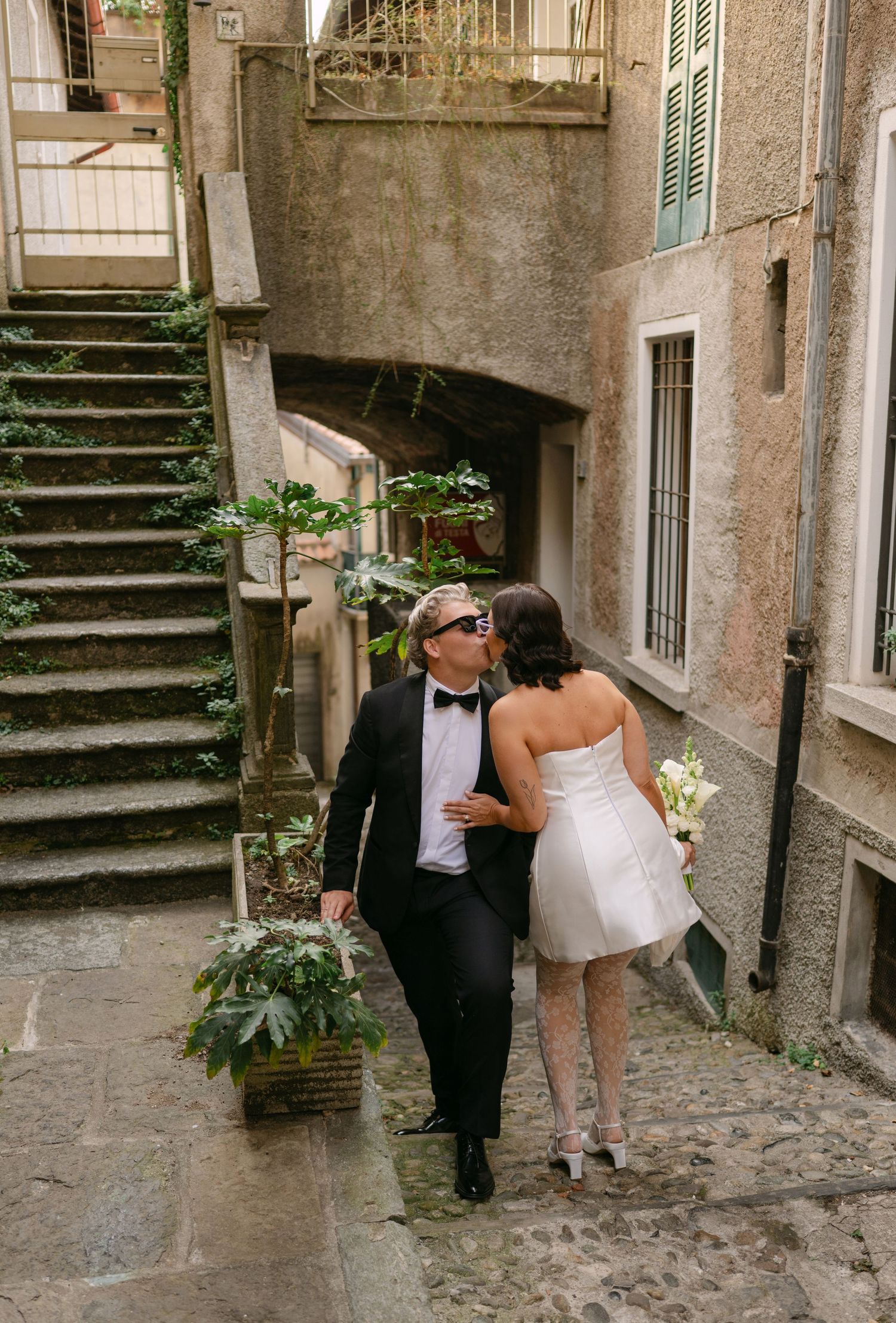 Couple in wedding attire kissing on cobblestone alley steps in a charming European village setting.