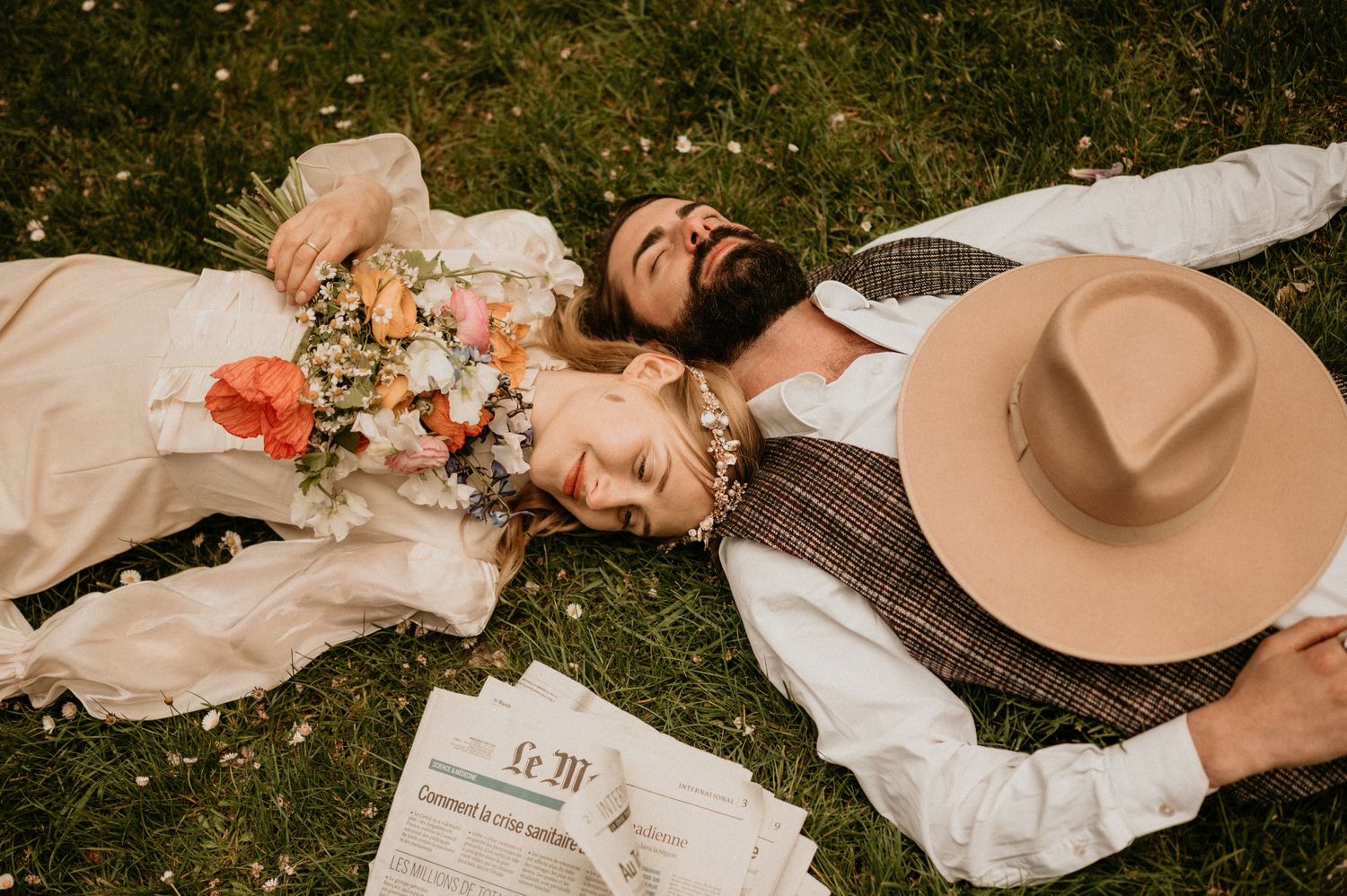 A couple resting on grass with a bouquet and tan hat, photographed from above in warm tones.