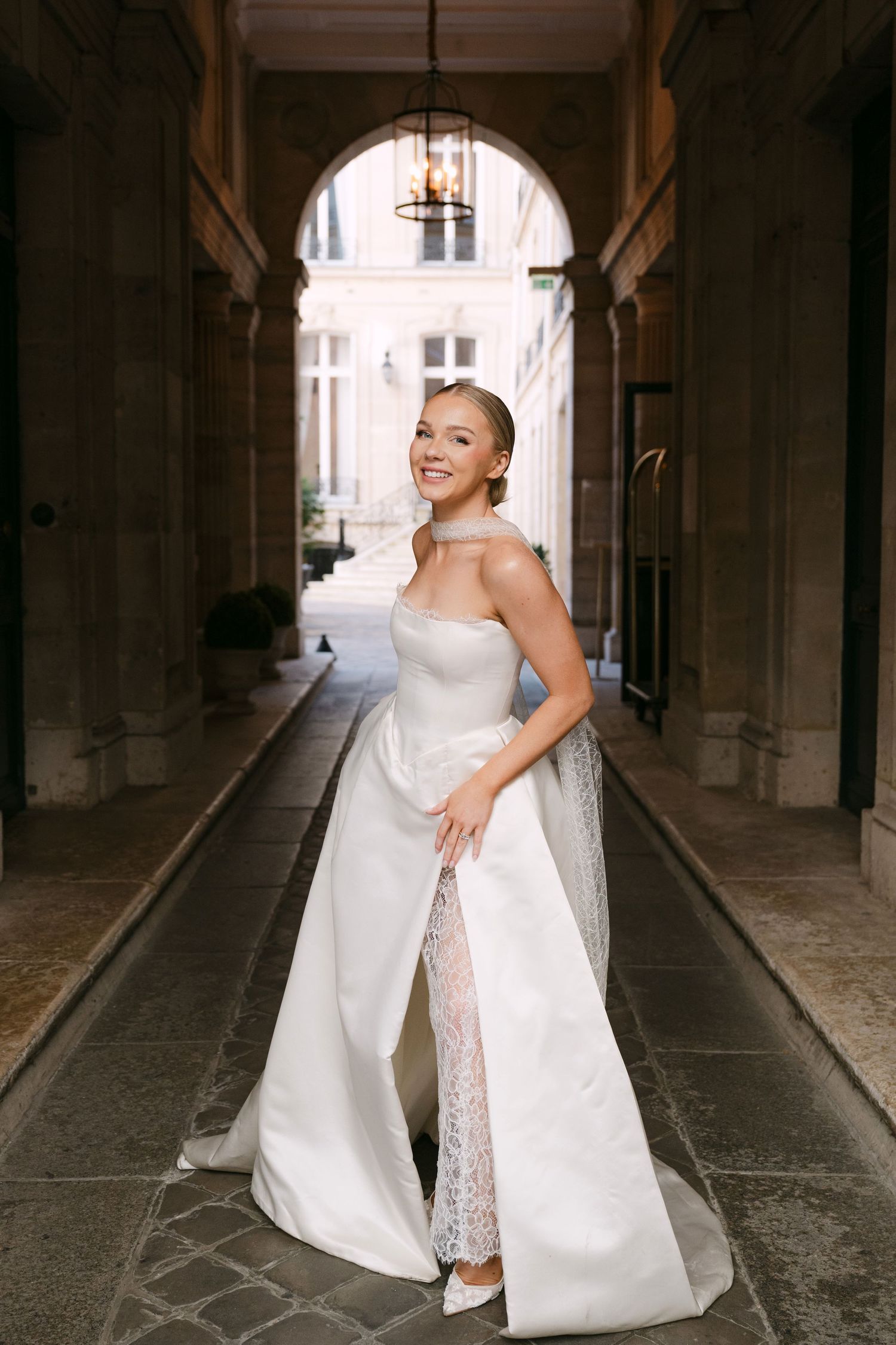 A bride in a white wedding gown twirls joyfully in an arched stone passageway.