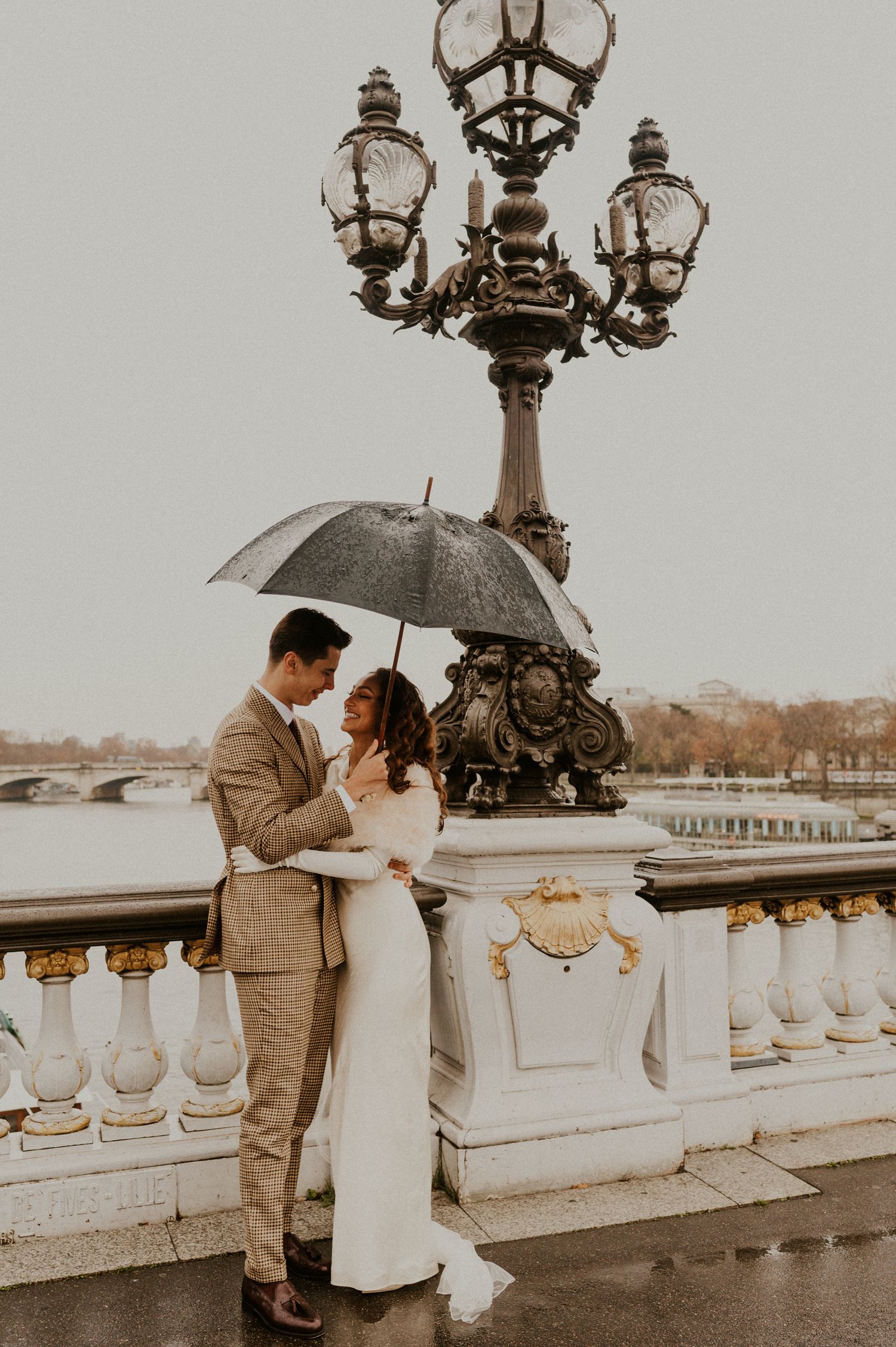 Romantic couple embracing under umbrella by vintage street lamp on Parisian bridge.