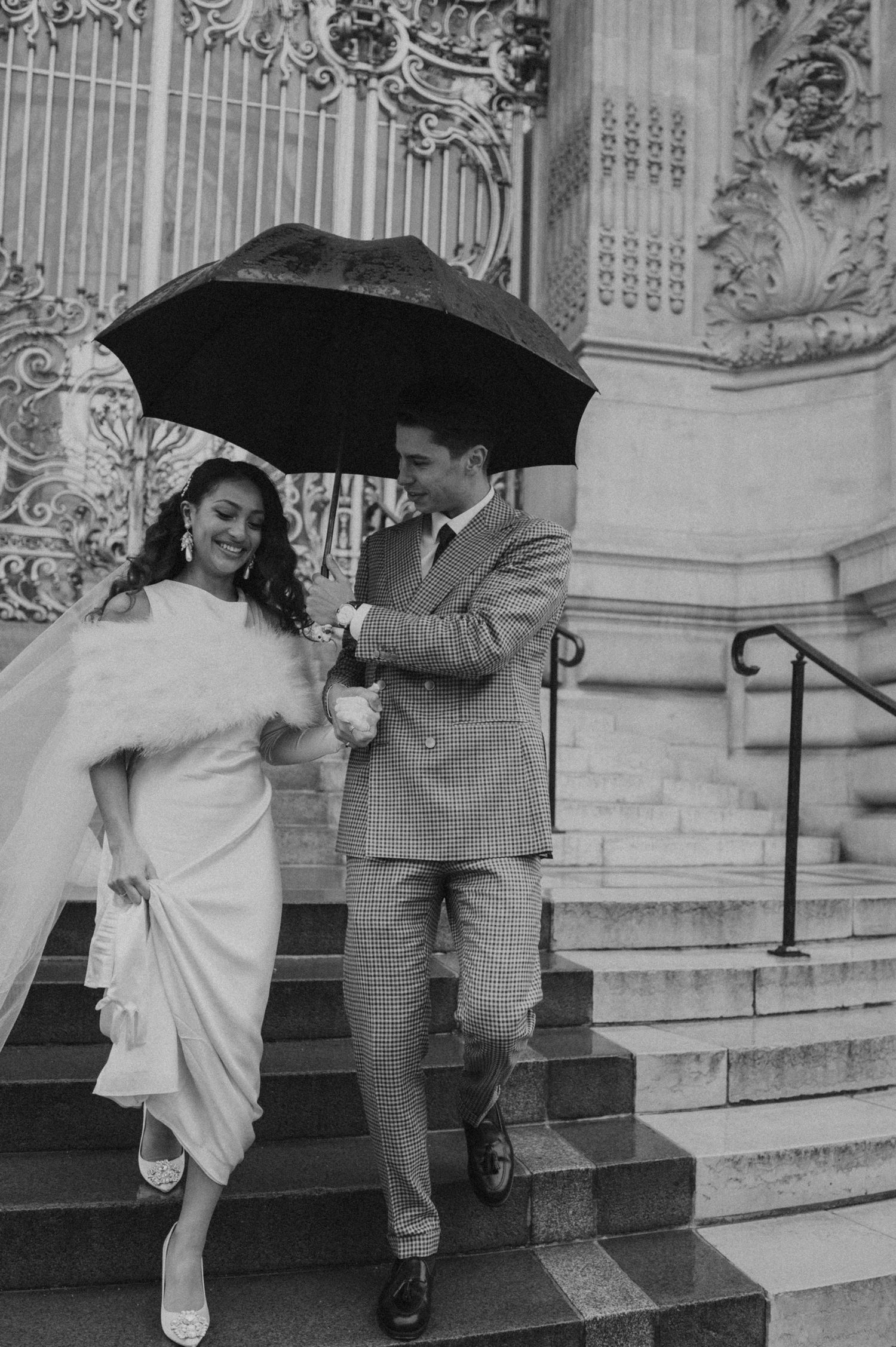 Black and white photo of a romantic couple on grand steps sharing an umbrella in vintage wedding attire.