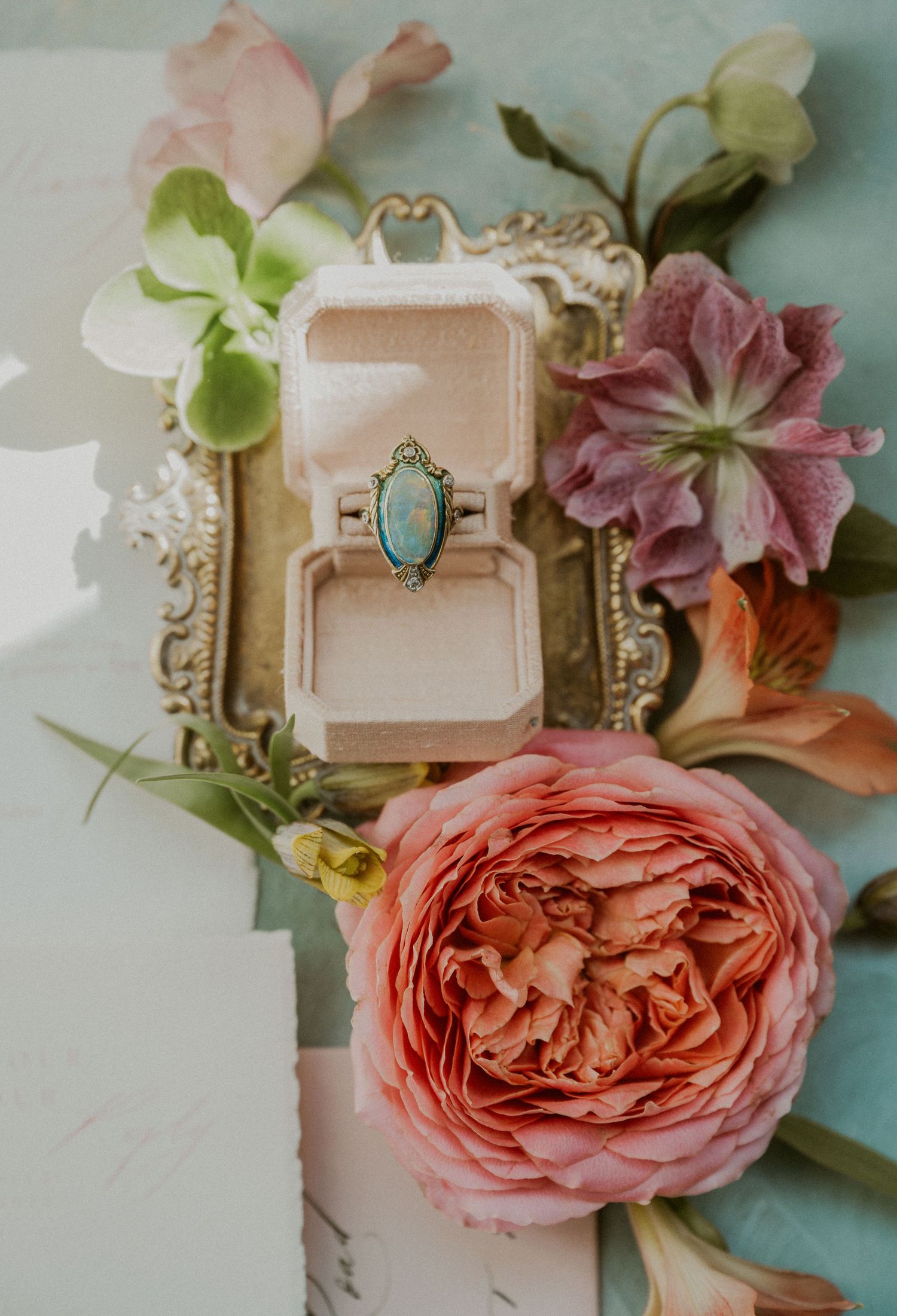 Vintage opal engagement ring in velvet box surrounded by pink garden roses and purple flowers on teal background.