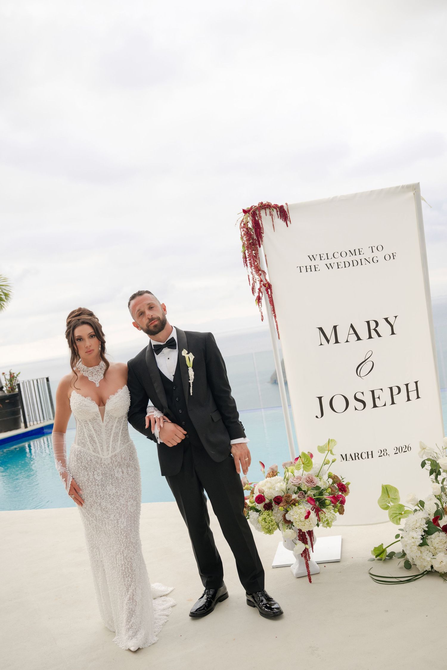 Mary and Joseph wedding welcome sign with bride in lace gown and groom in tuxedo by pool.