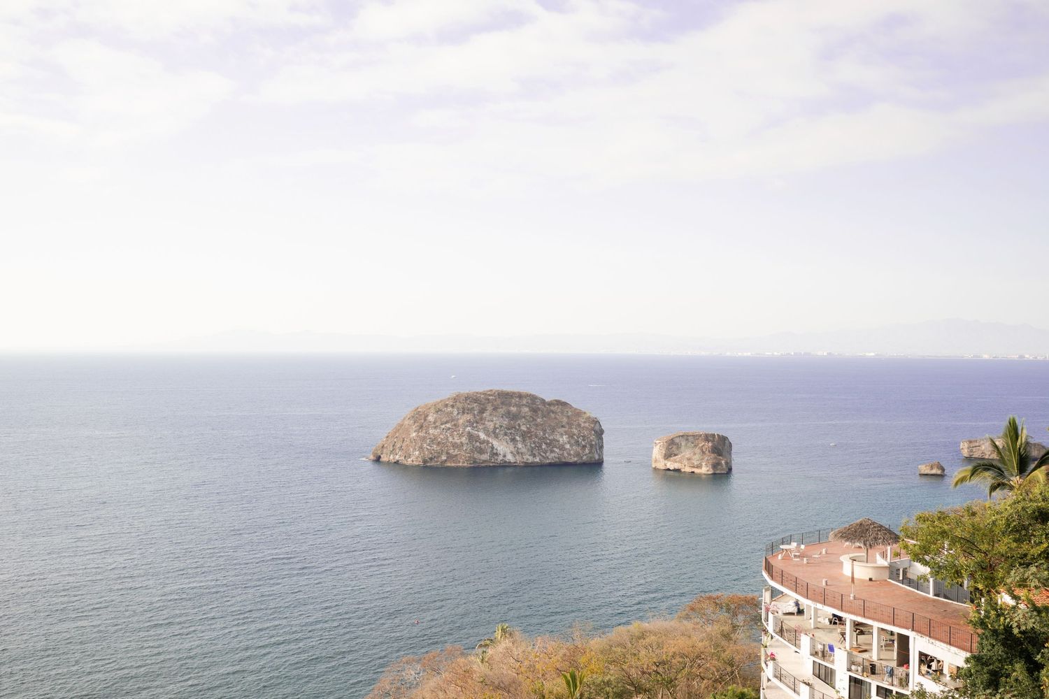 Two rocky islands rise from calm blue ocean waters near a coastal building with red roof in Puerto Vallarta, Mexico.
