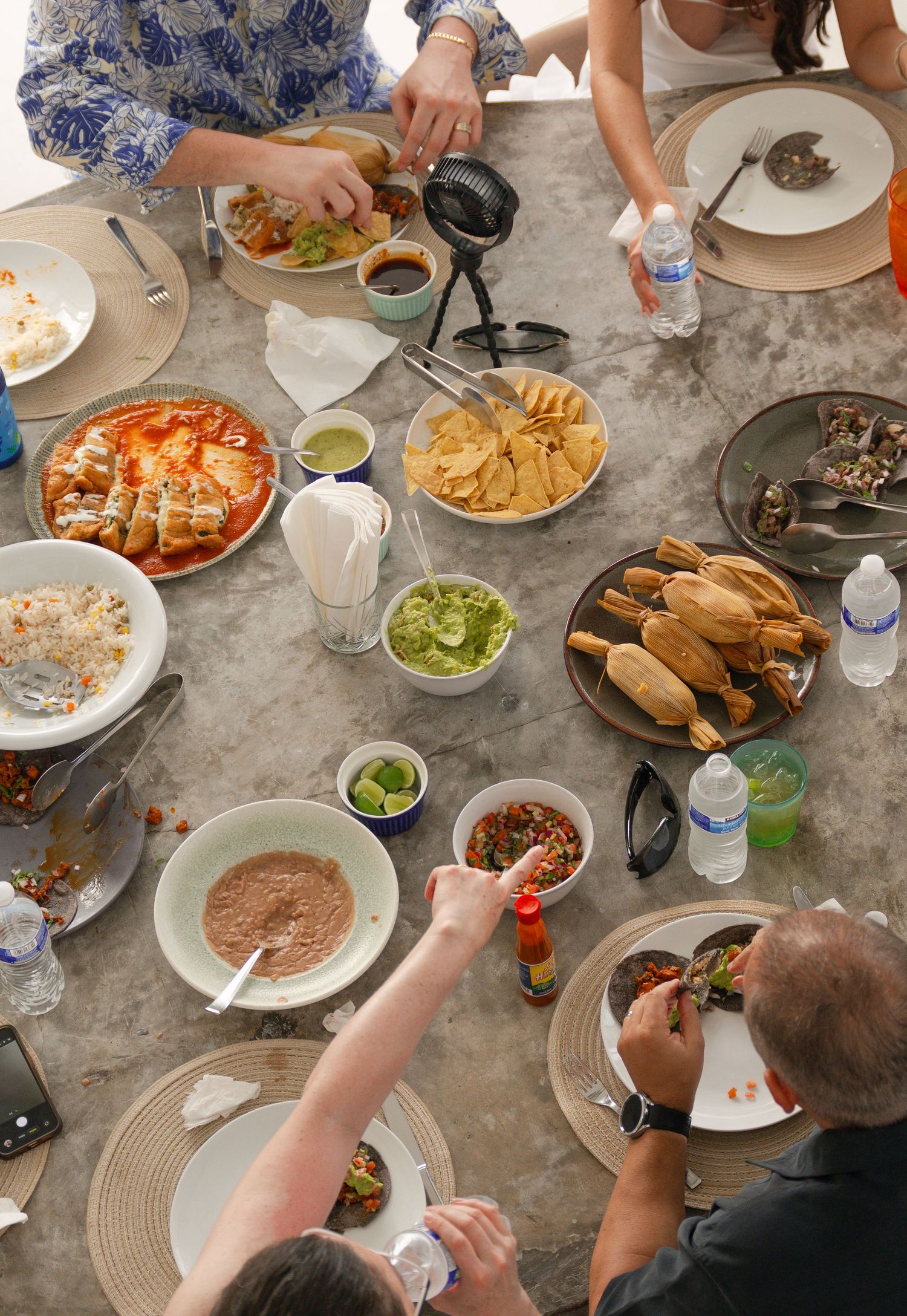 Overhead view of a Mexican feast with guacamole, chips, tamales, refried beans, and salsa shared among friends.