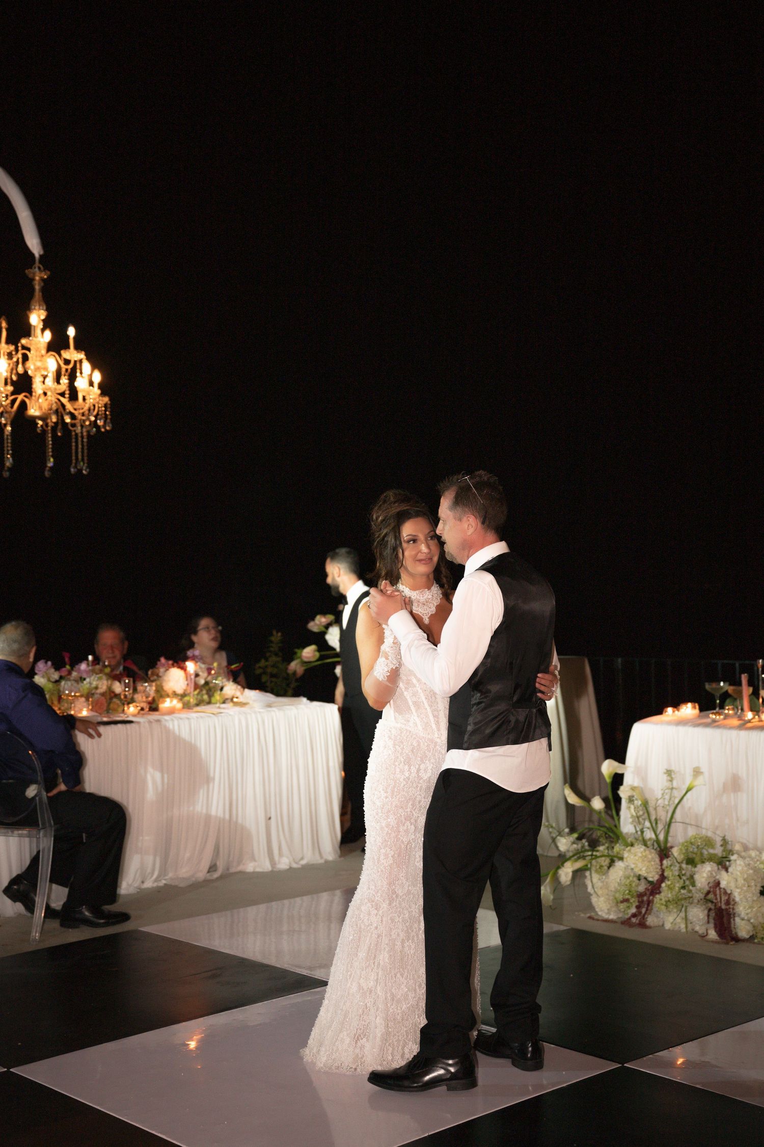 Newlyweds share romantic first dance on black and white floor at candlelit outdoor wedding.