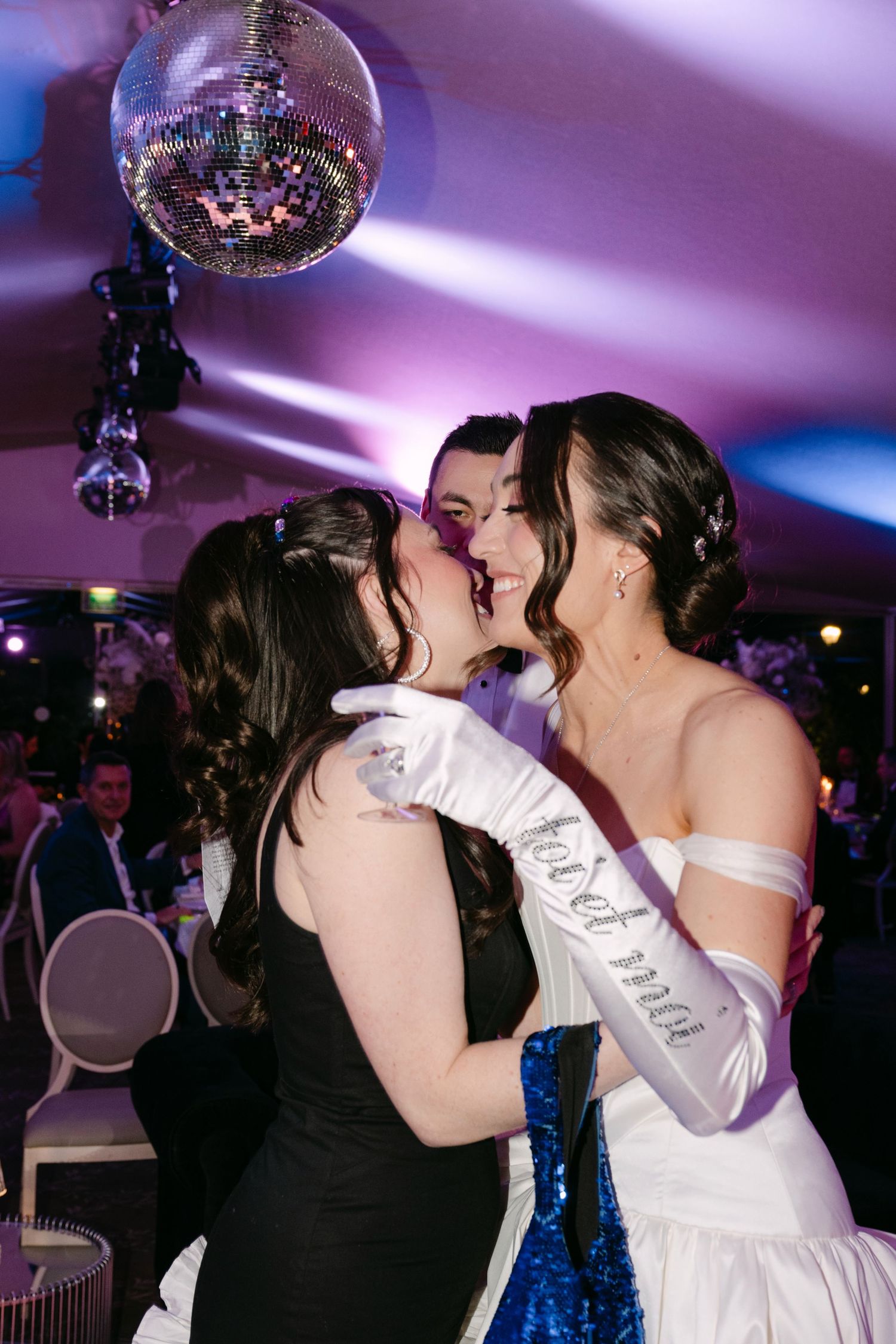 Bride in white gown with satin gloves shares kiss with female guest at wedding reception.