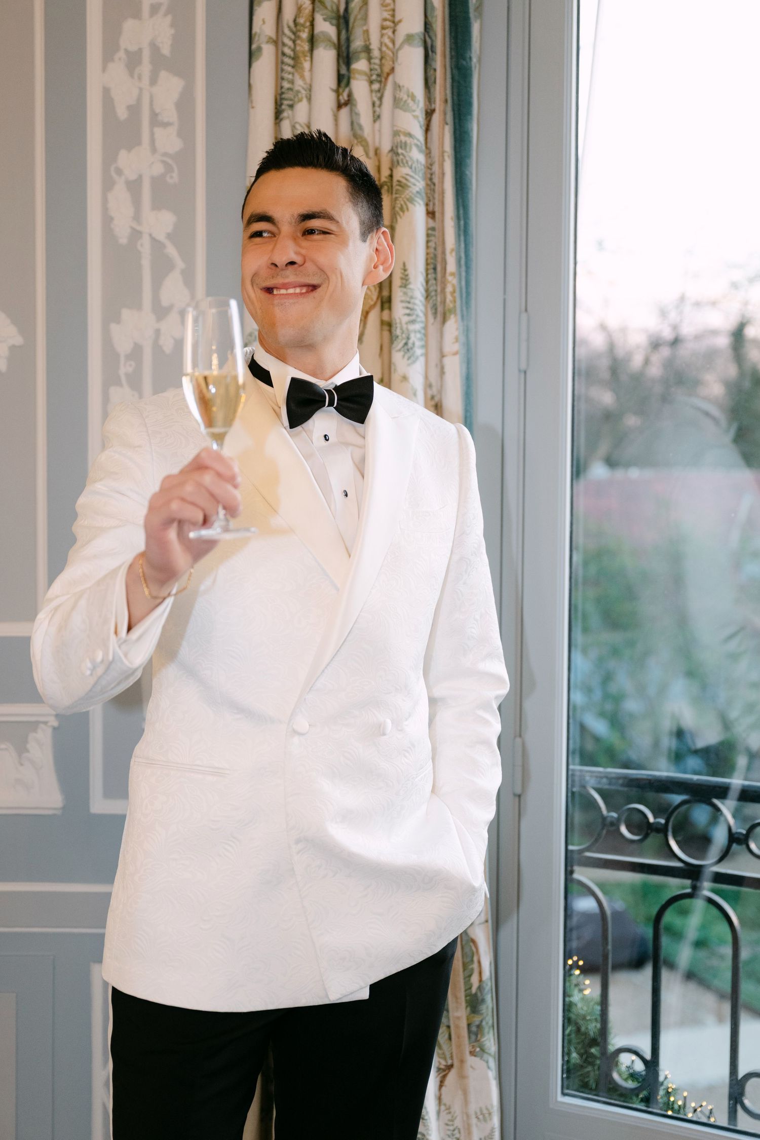 Groom in white double-breasted tuxedo jacket holding champagne glass near window at wedding.