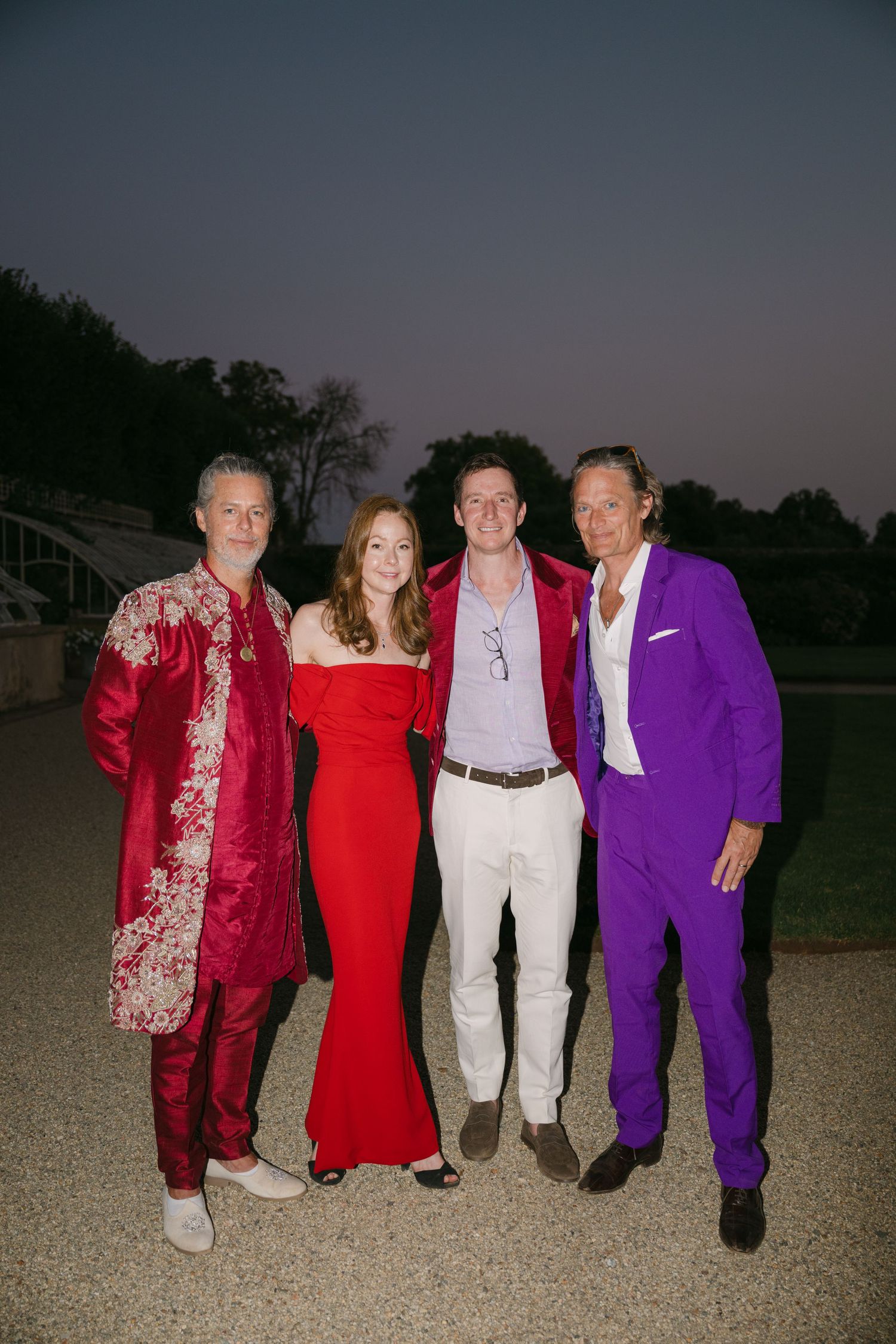 Four elegantly dressed wedding guests posing outdoors at dusk in vibrant red and purple formal attire.
