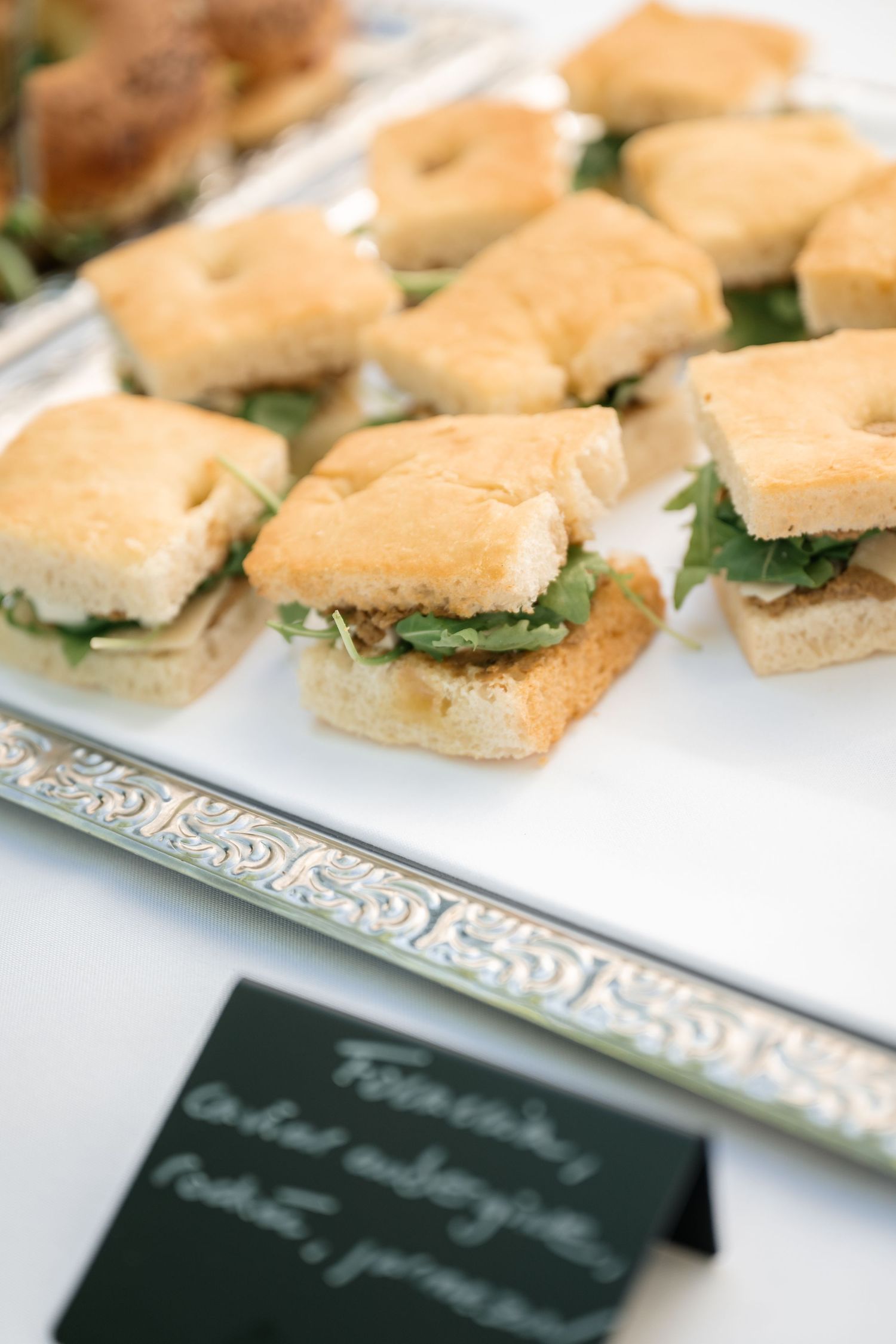 Tray of focaccia sandwiches with greens and cheese displayed at a catered event.