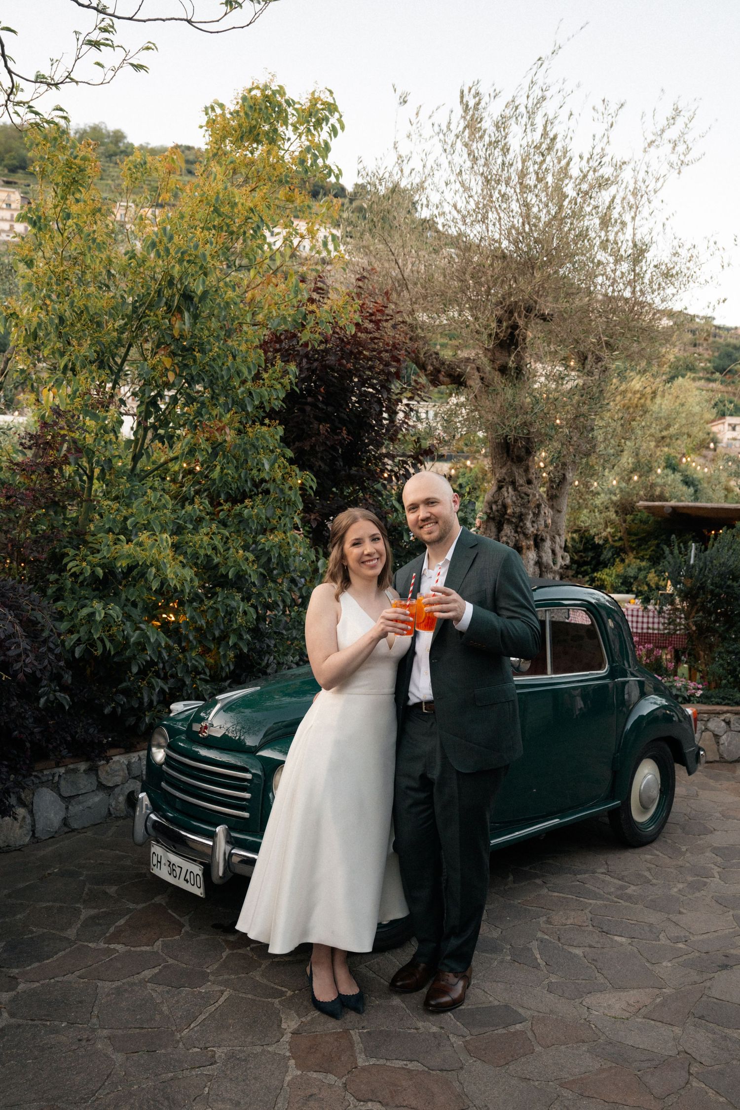 Happy wedding couple toasting with Aperol Spritz beside a vintage green Fiat car outdoors.
