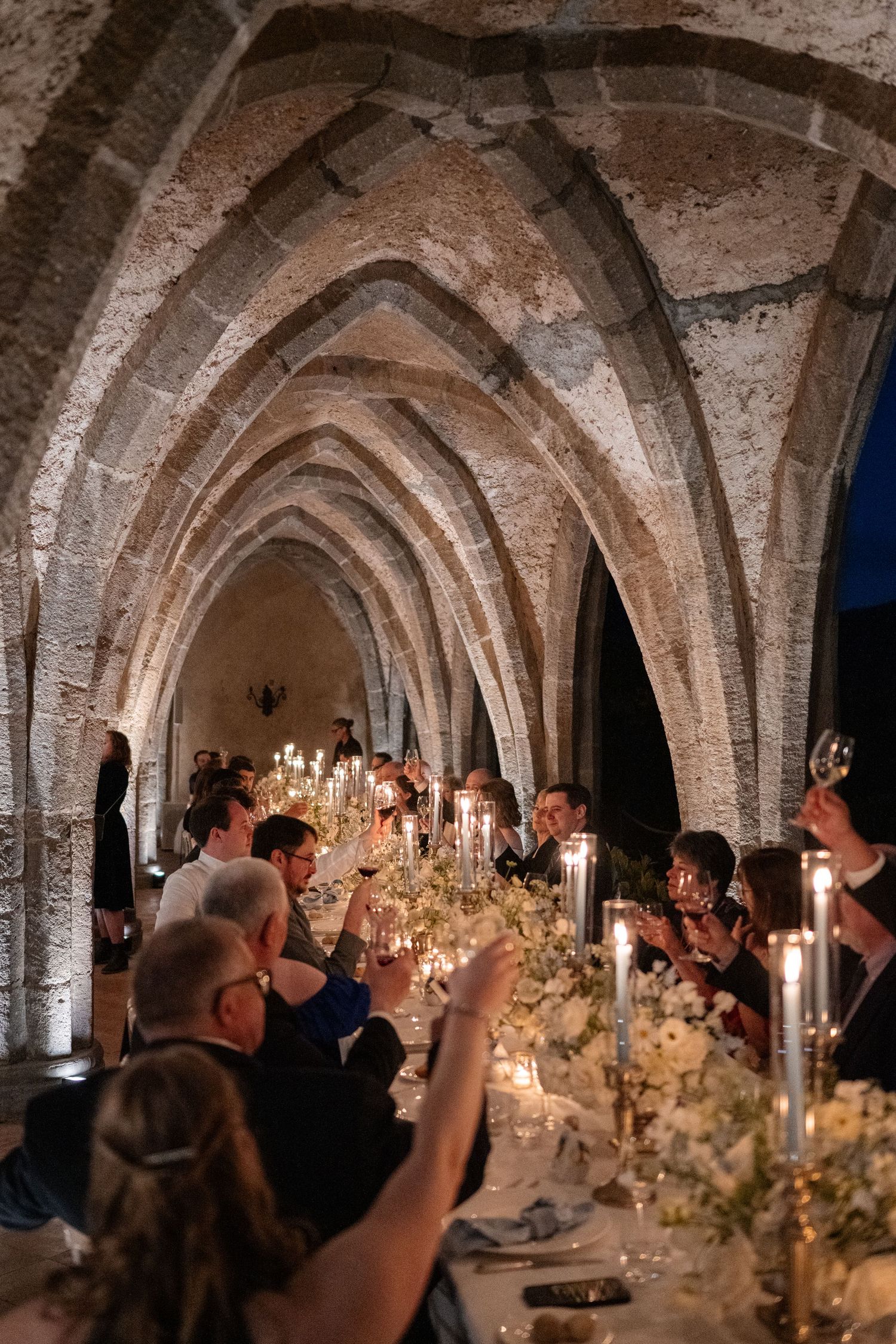 Wedding guests raise glasses in a toast during a candlelit dinner under dramatic Gothic stone arches at night.