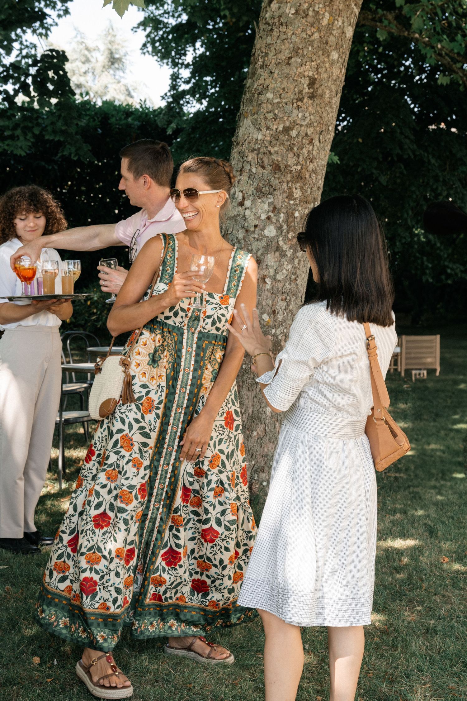 Guests in summer dresses socializing under trees at an elegant outdoor garden wedding cocktail hour.