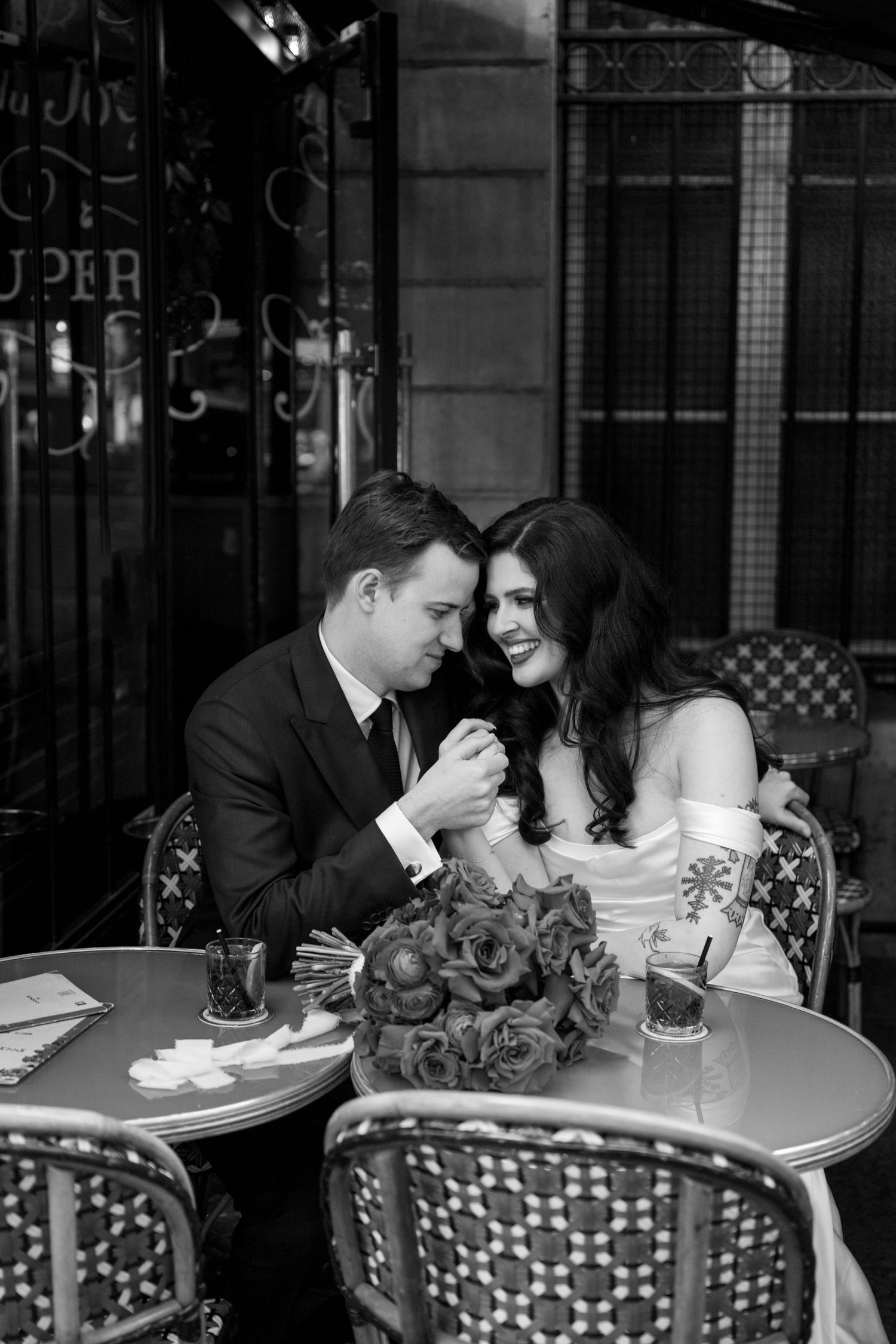Black and white photo of smiling bride and groom sharing intimate moment at Parisian cafe table.