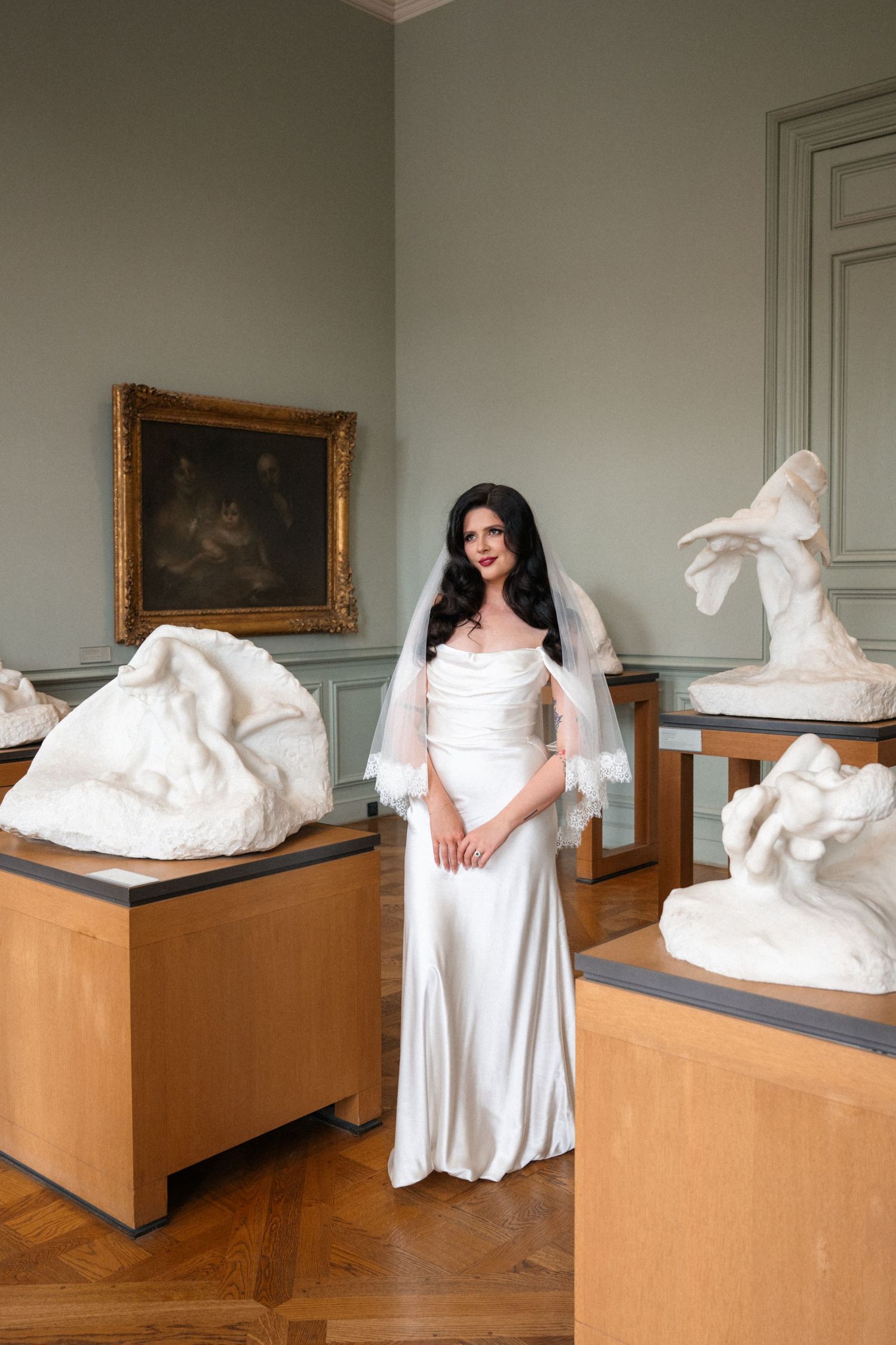 Elegant bride in white satin gown and lace veil poses among classical sculptures in a museum gallery.