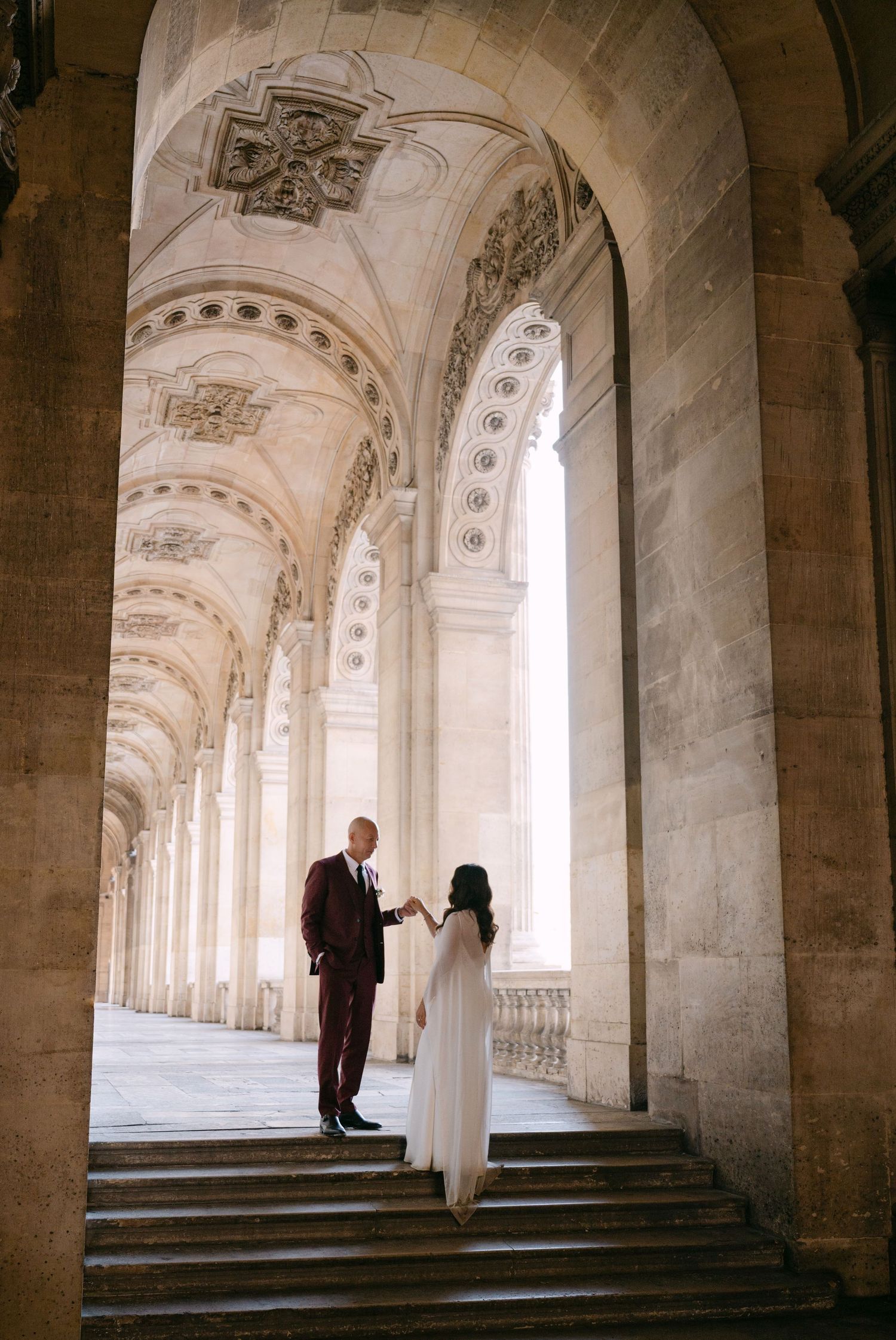Bride and groom share intimate moment on steps beneath ornate arched colonnade at the Louvre Paris.