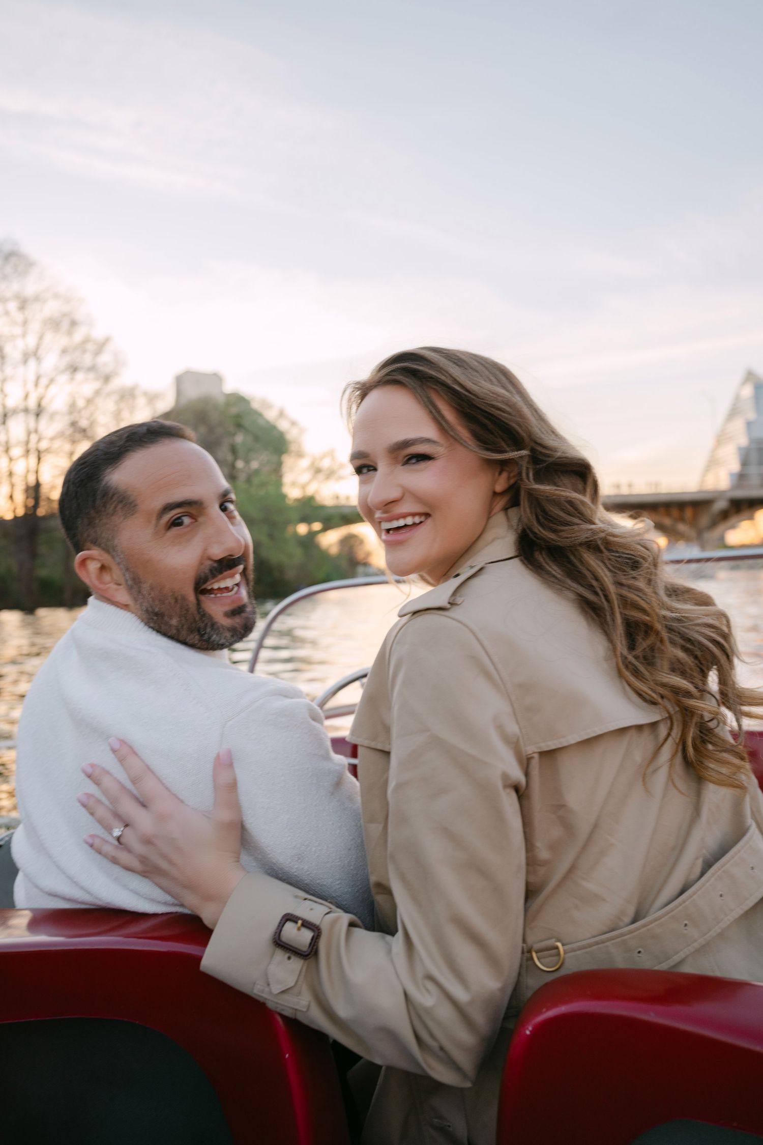 Laughing couple embracing on a red boat at sunset, woman in trench coat showing engagement ring on her hand.