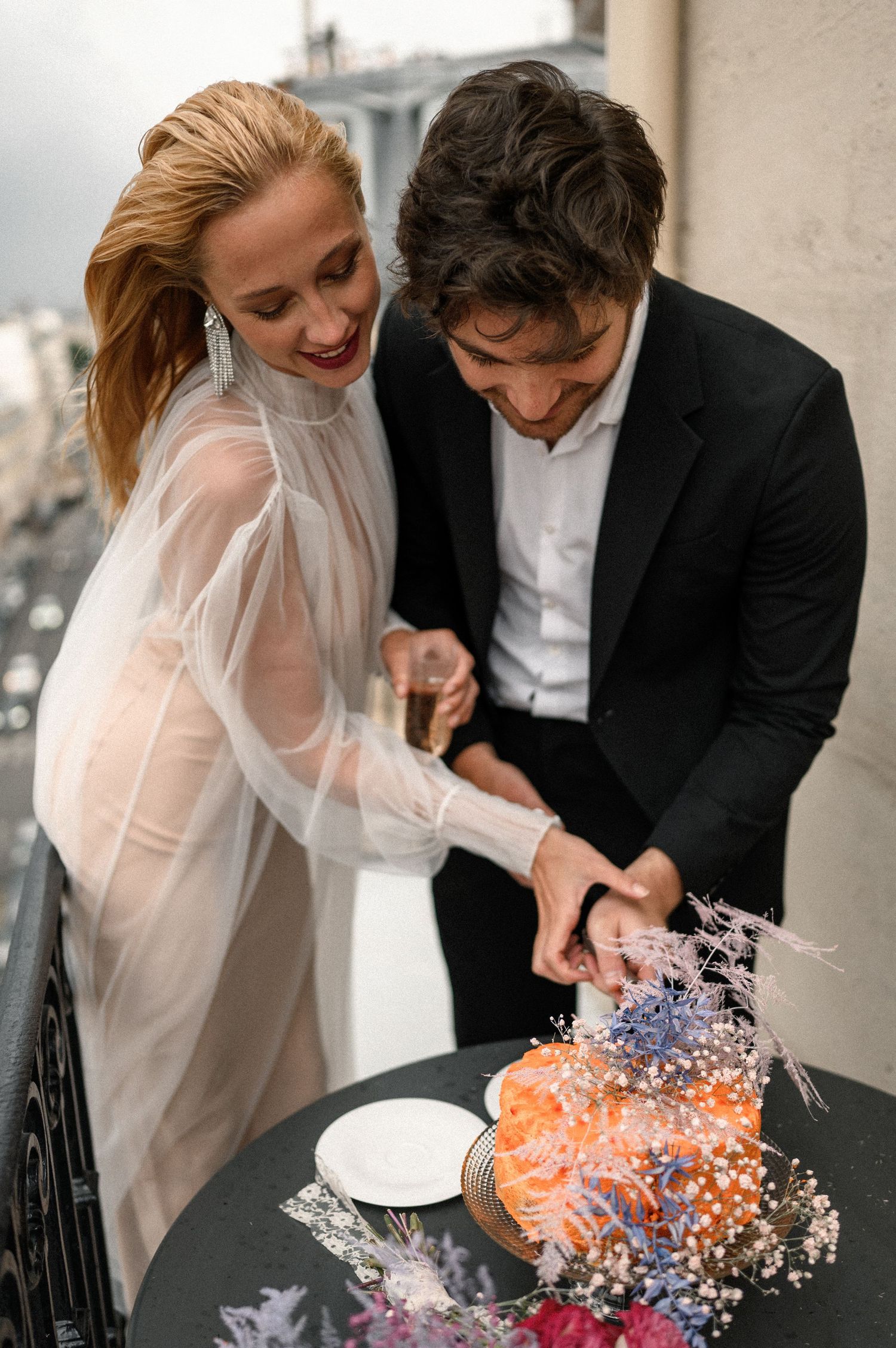 Bride and groom cutting a vibrant orange wedding cake adorned with dried flowers on a Parisian balcony.