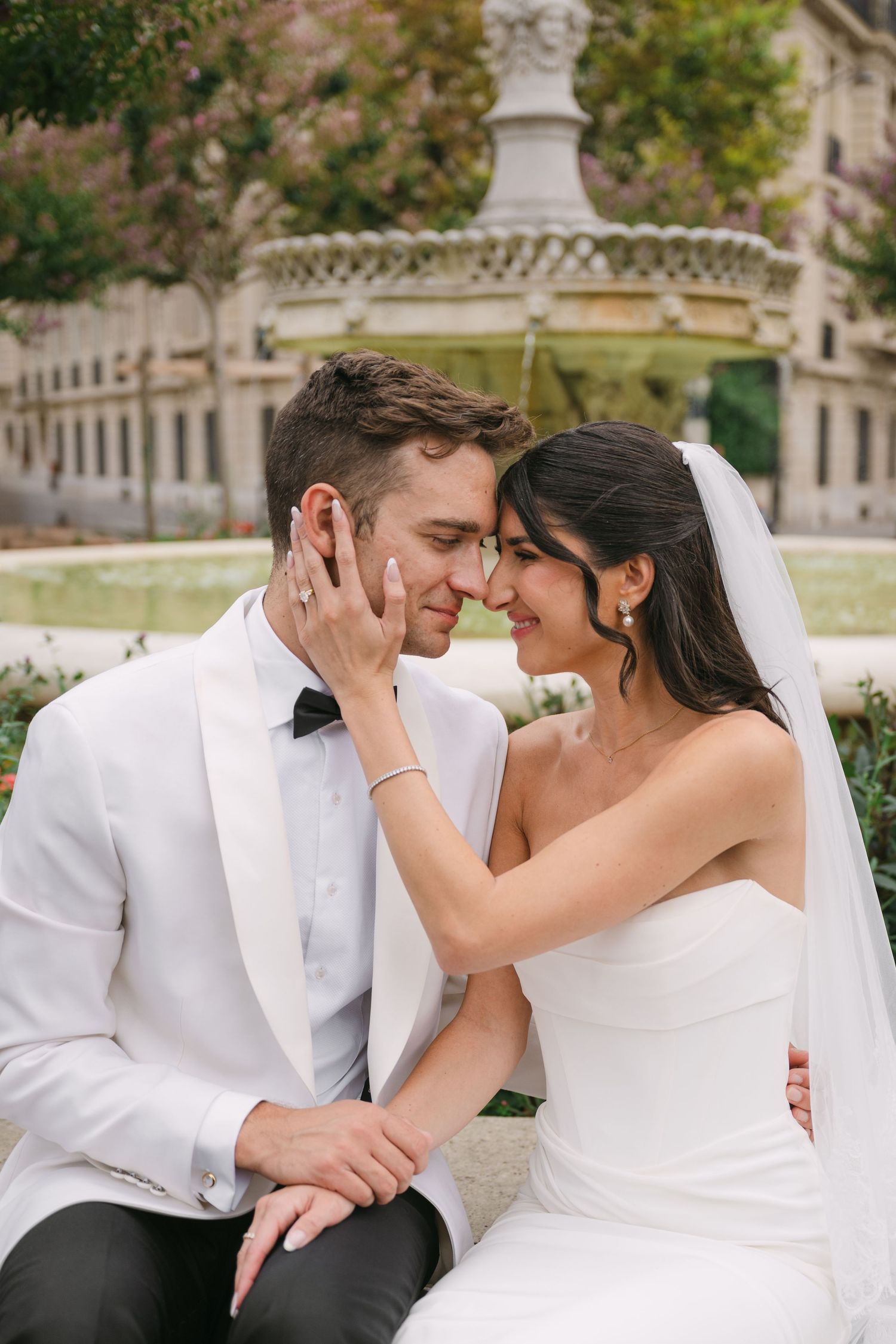Smiling bride and groom sit together by a fountain in a Paris garden, touching foreheads tenderly.